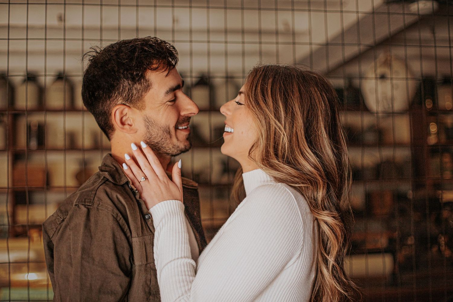 A smiling couple shares a romantic moment, the woman tenderly touching the mans face while nearly kissing indoors.