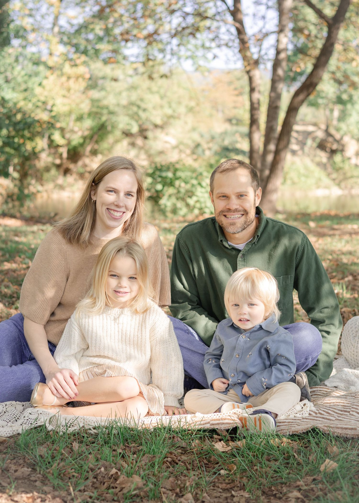 Family of four smiling outdoors on a blanket in a park during fall, with two young children in front.