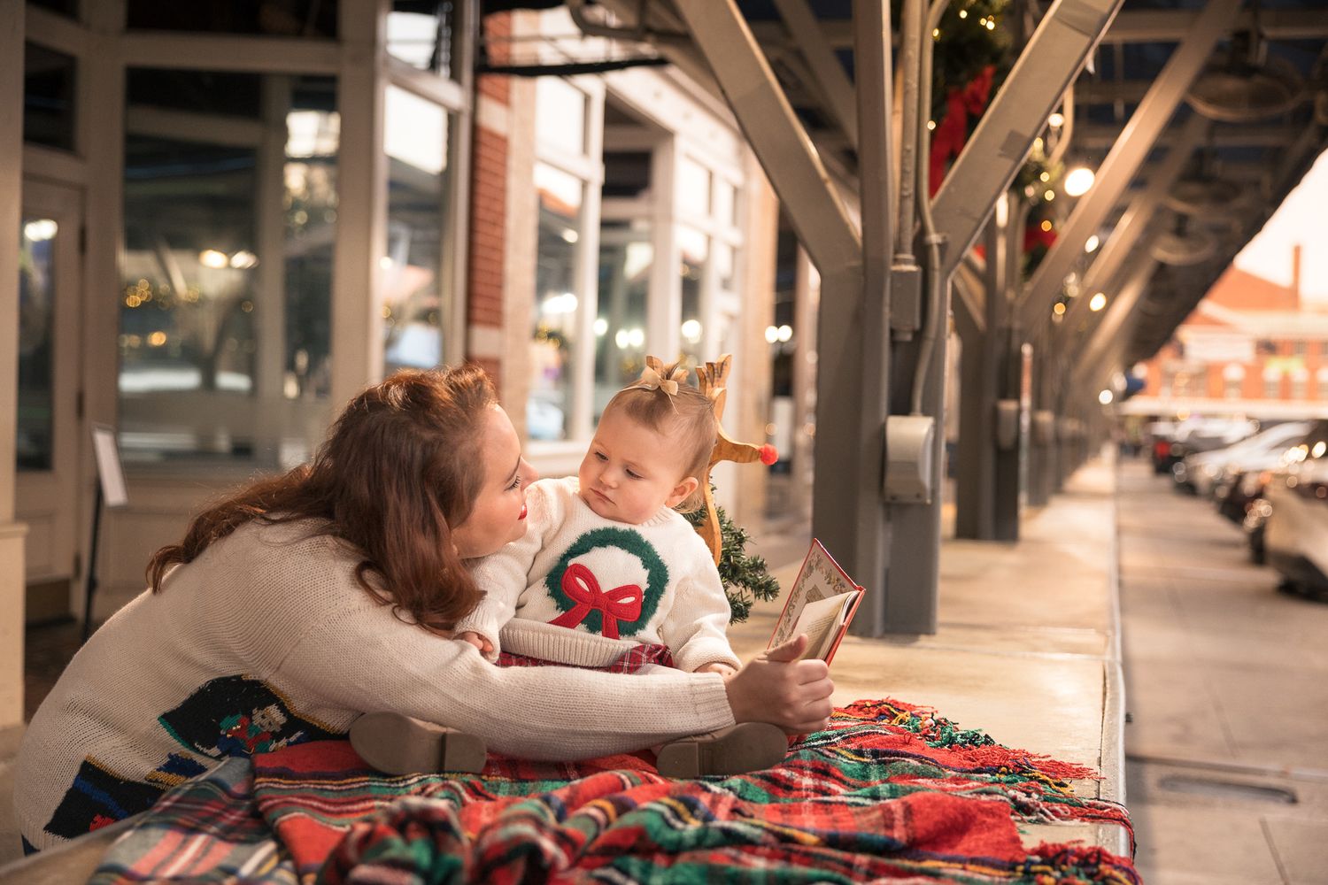 Mother and baby girl lying on plaid blanket looking at phone together outdoors during Christmas holiday season.