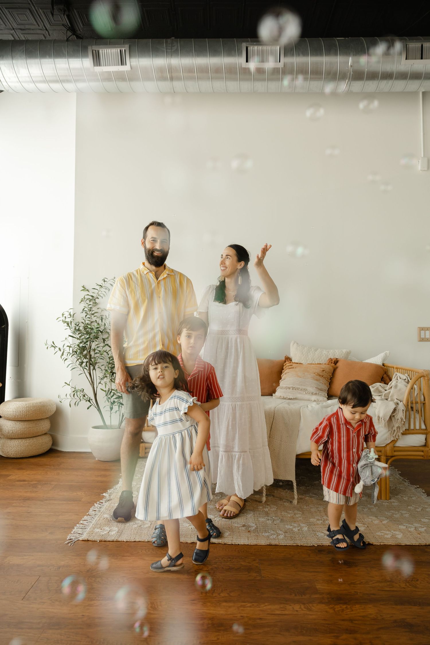Family of five playing with bubbles indoors, featuring a bearded dad, mom in white dress, and three young children on hardwood floors.