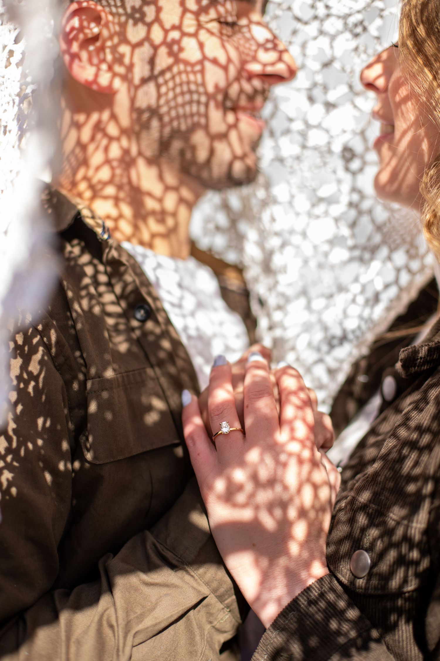 Couple holding hands with engagement ring visible, dappled sunlight creating lace patterns on their faces and olive jackets.