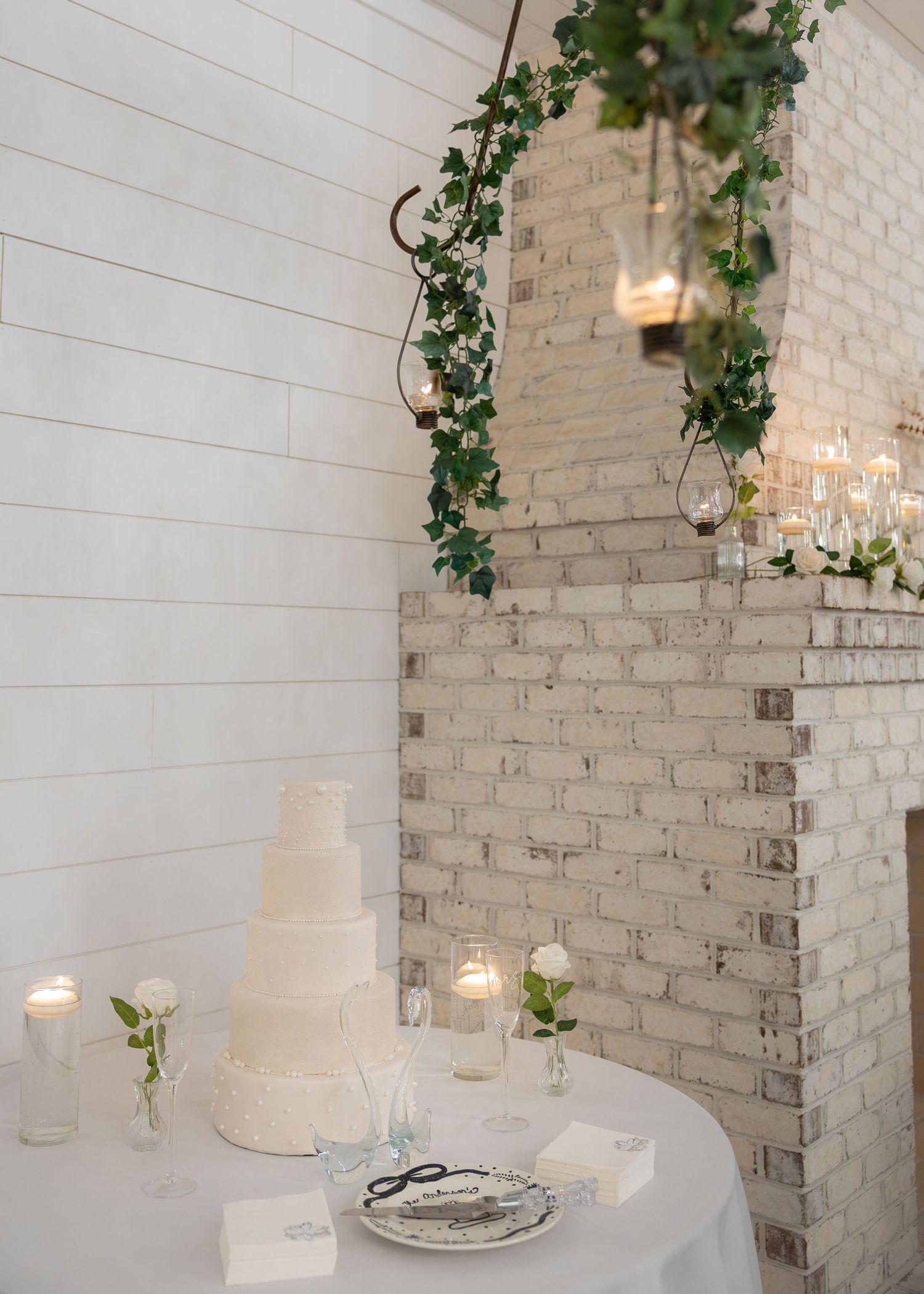 Elegant white wedding cake on draped table with candles, roses, and ivy garland against whitewashed brick wall.