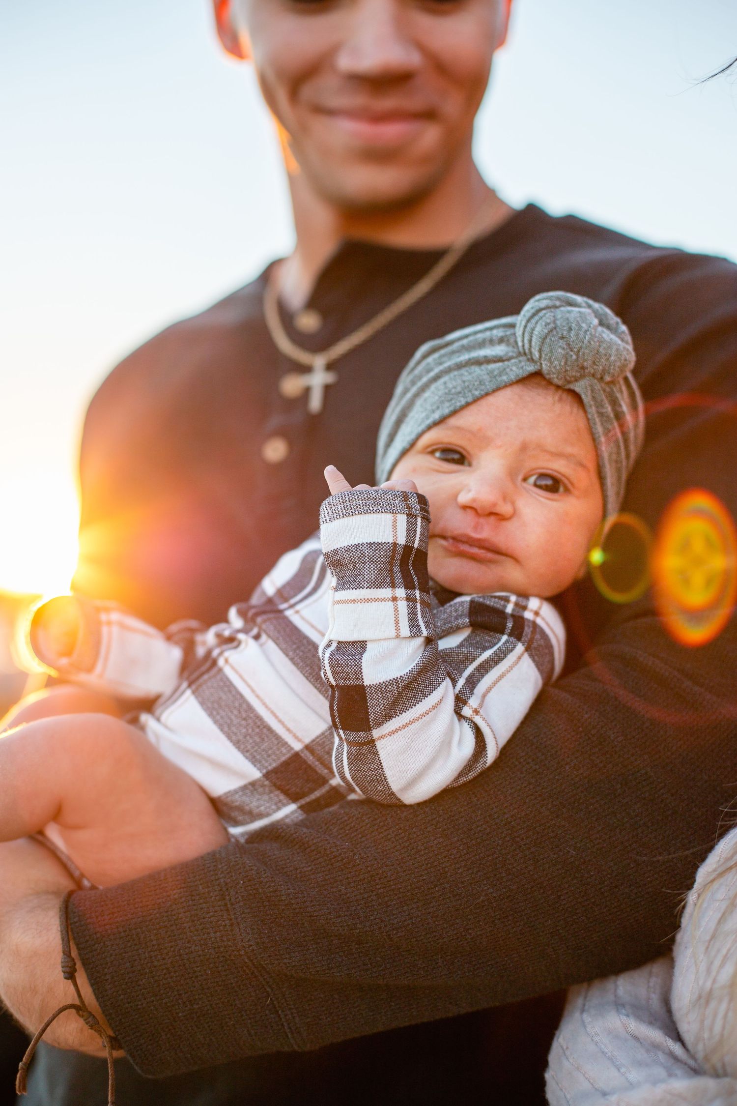 Young man holding a newborn baby in a plaid onesie and gray turban headband, bathed in golden sunset light.