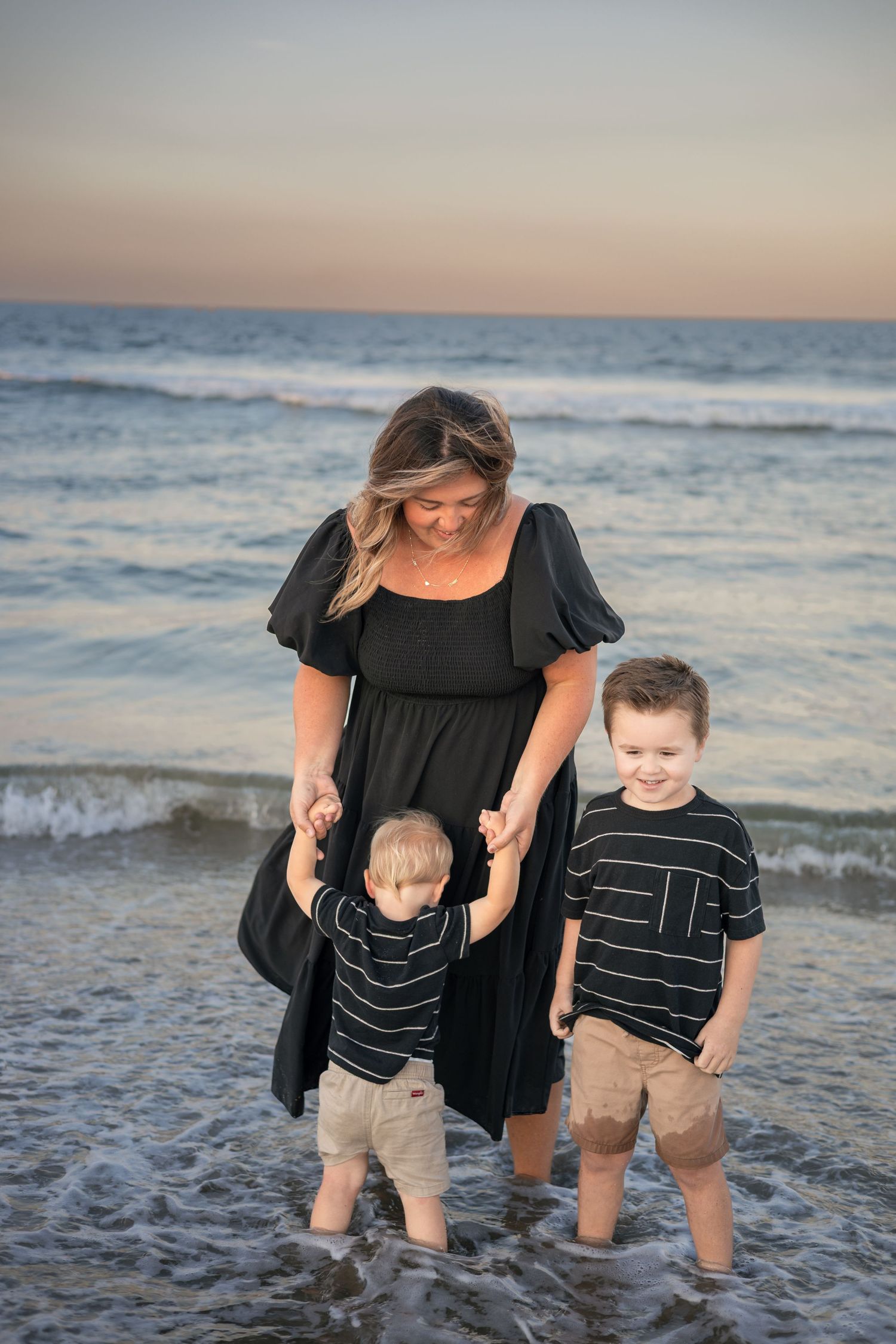A mother in a black dress holds hands with her two young sons while standing in ocean waves at sunset on the beach.