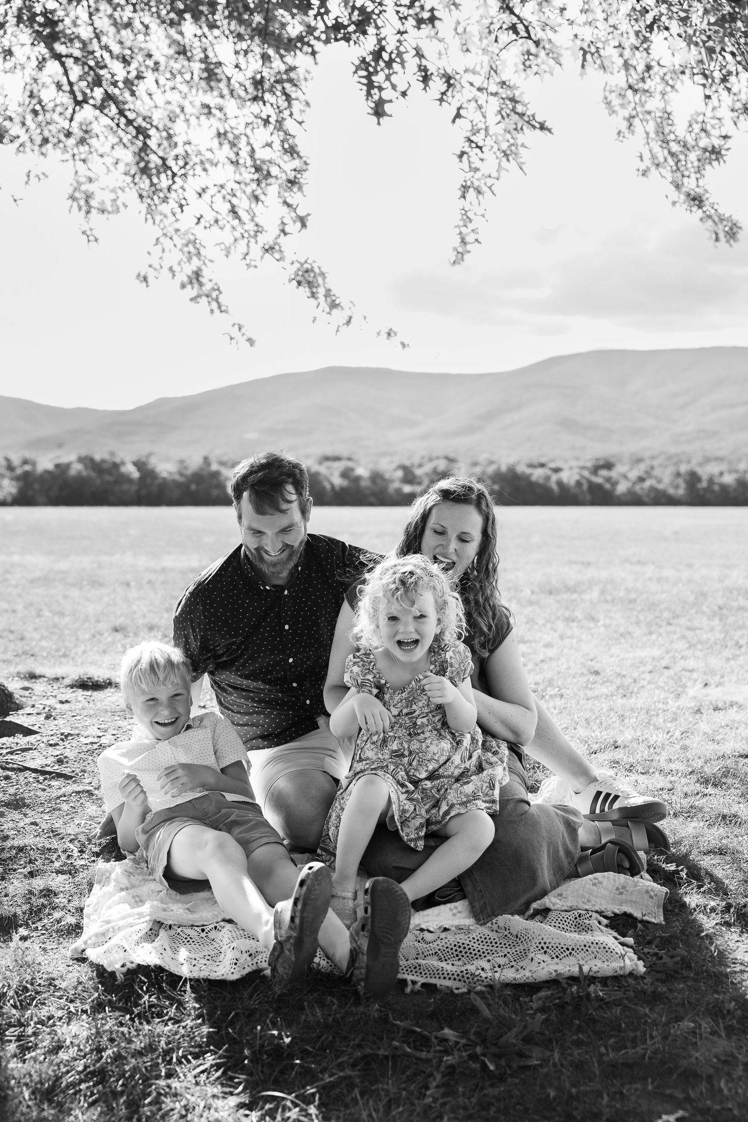 Black and white family photo of parents and two young children sitting on a blanket under a tree with mountains in the background.