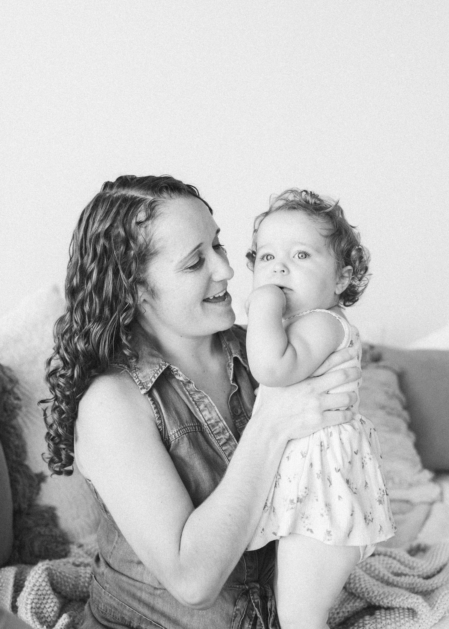 Black and white photo of a smiling mother holding her baby girl in a floral dress, sharing a tender moment.