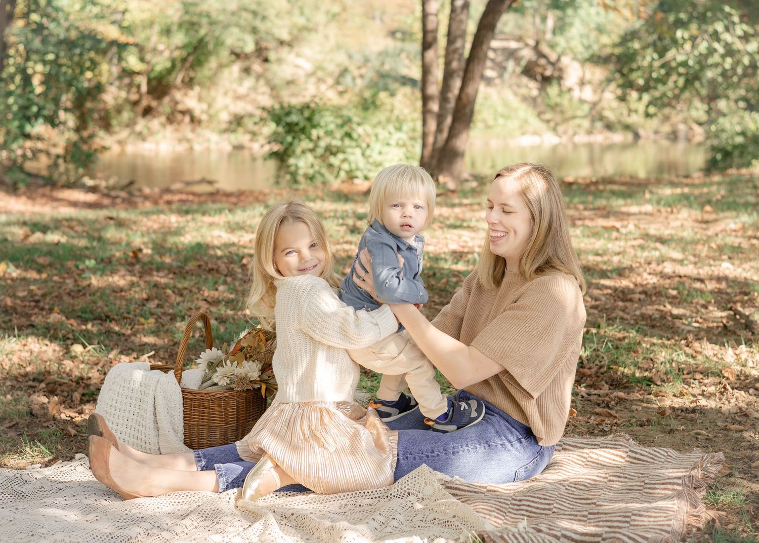 A mother sits on a blanket outdoors with two young children near a wicker basket in a sunlit park setting.