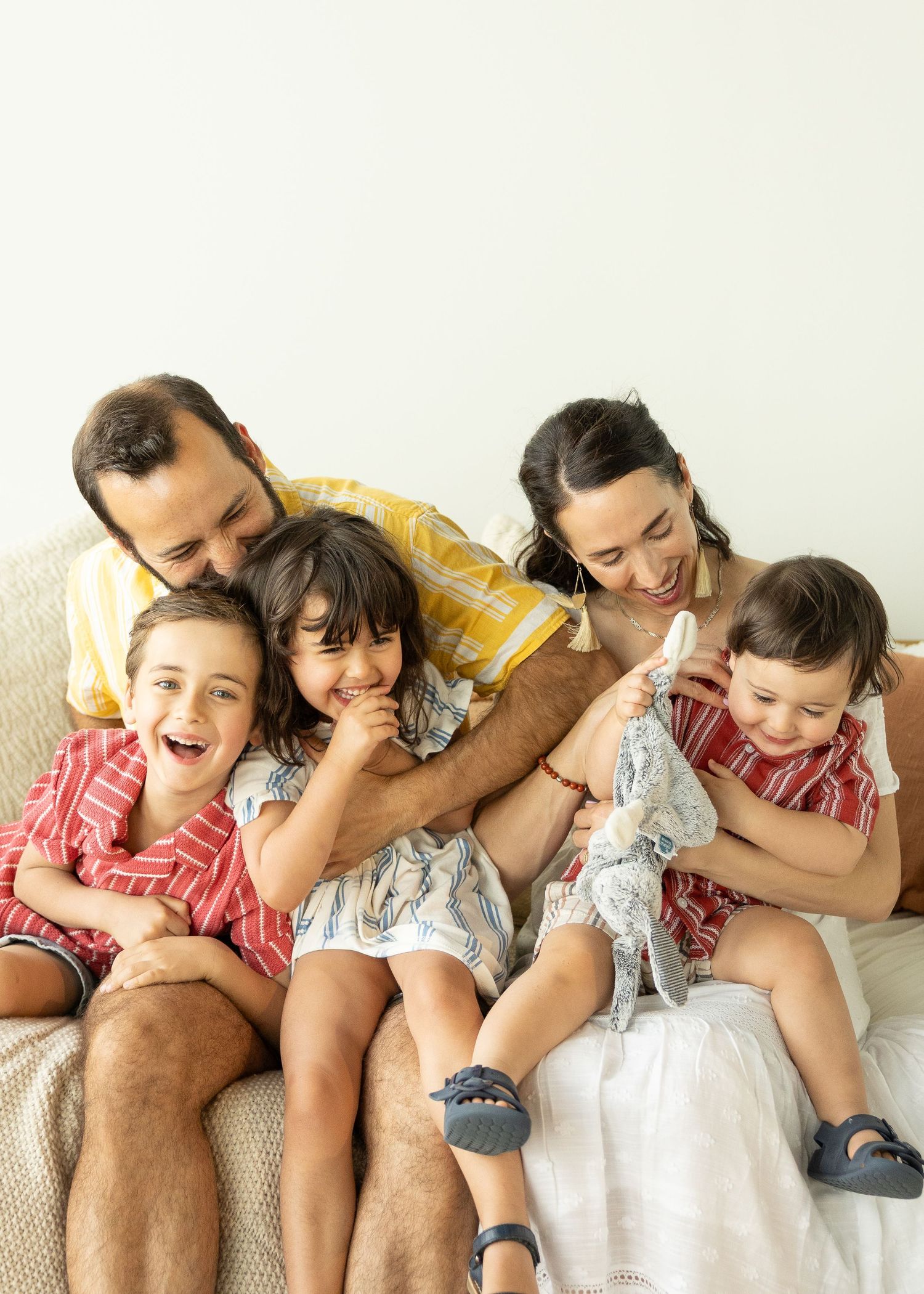 A joyful family of five sits together on a couch, laughing and cuddling, dressed in matching red and blue outfits.