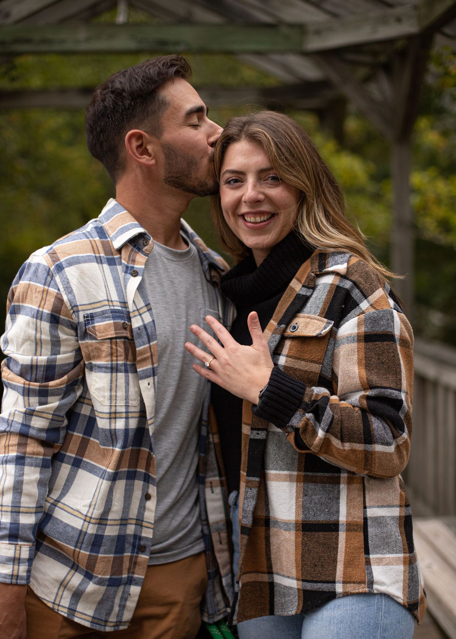 Couple in matching plaid shirts celebrating engagement, man kissing woman's cheek as she shows off her ring.