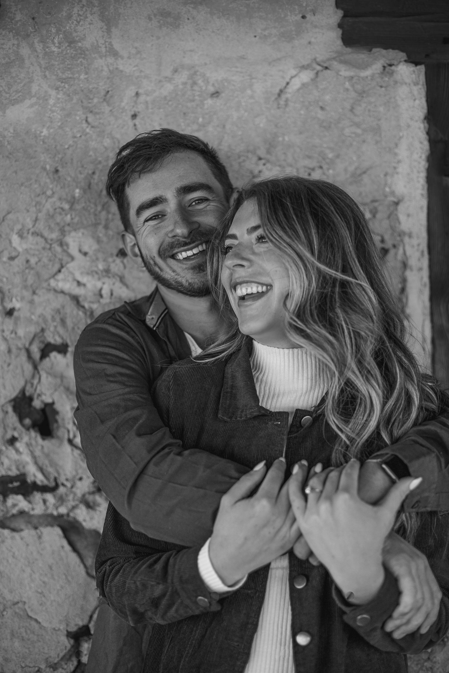 Black and white photo of a happy couple embracing and laughing together against a rustic stone wall.