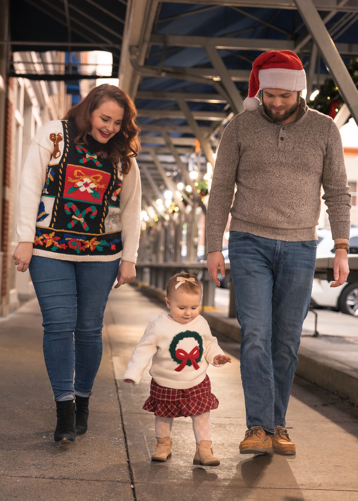 Family in Christmas sweaters walks downtown, toddler in wreath sweater holds parents hands, man wears Santa hat.