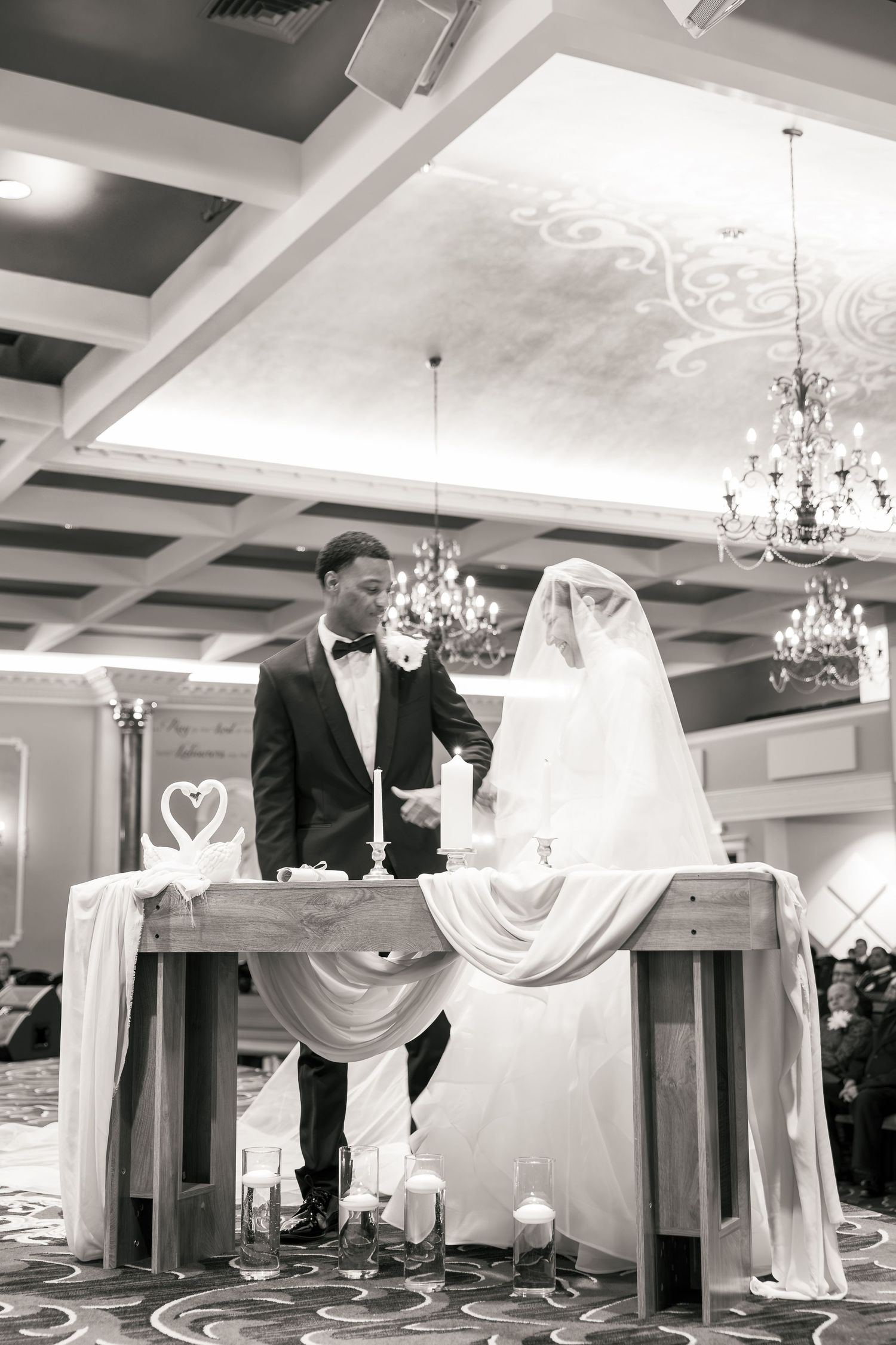 Black and white photo of a bride and groom standing at altar table during wedding ceremony in elegant ballroom.