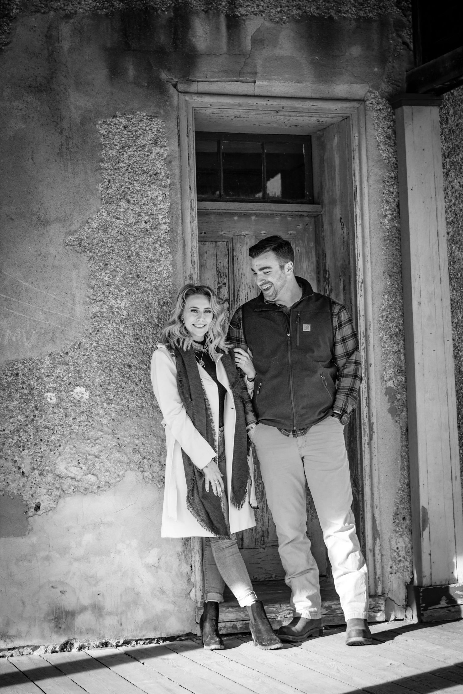 Black and white photo of a smiling couple standing in a rustic doorway, wearing casual winter clothing.