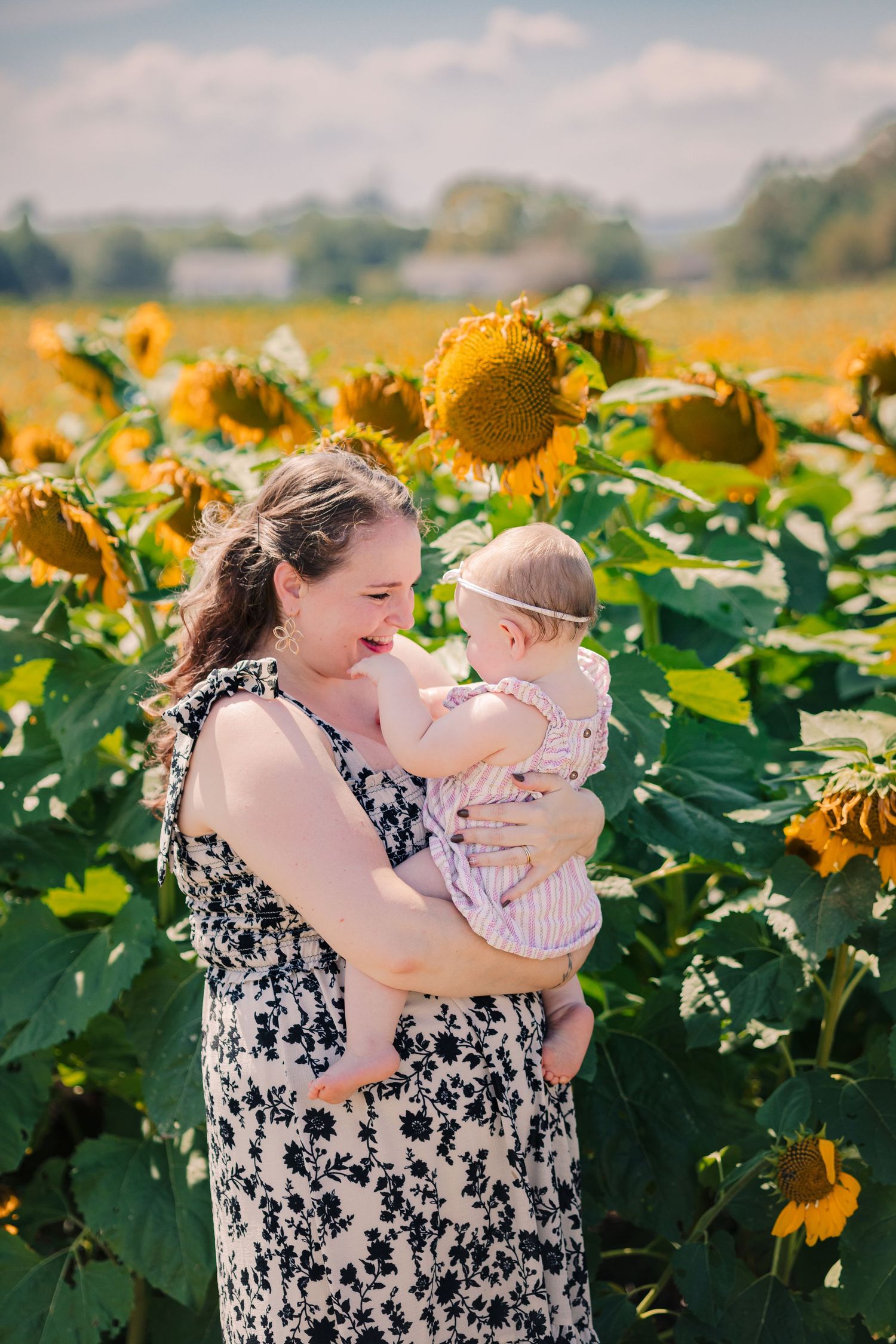 A mother smiles while holding her baby in a vibrant sunflower field on a sunny day. buchanan sunflower festival