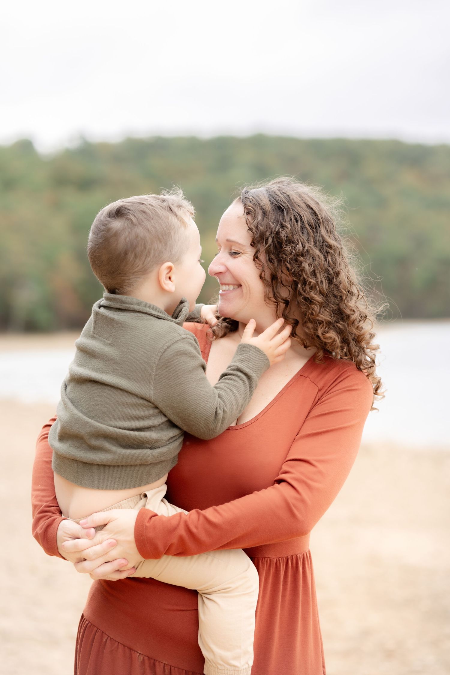 Smiling curly-haired woman in rust dress holding a toddler boy at a sandy lakeside on an overcast day.