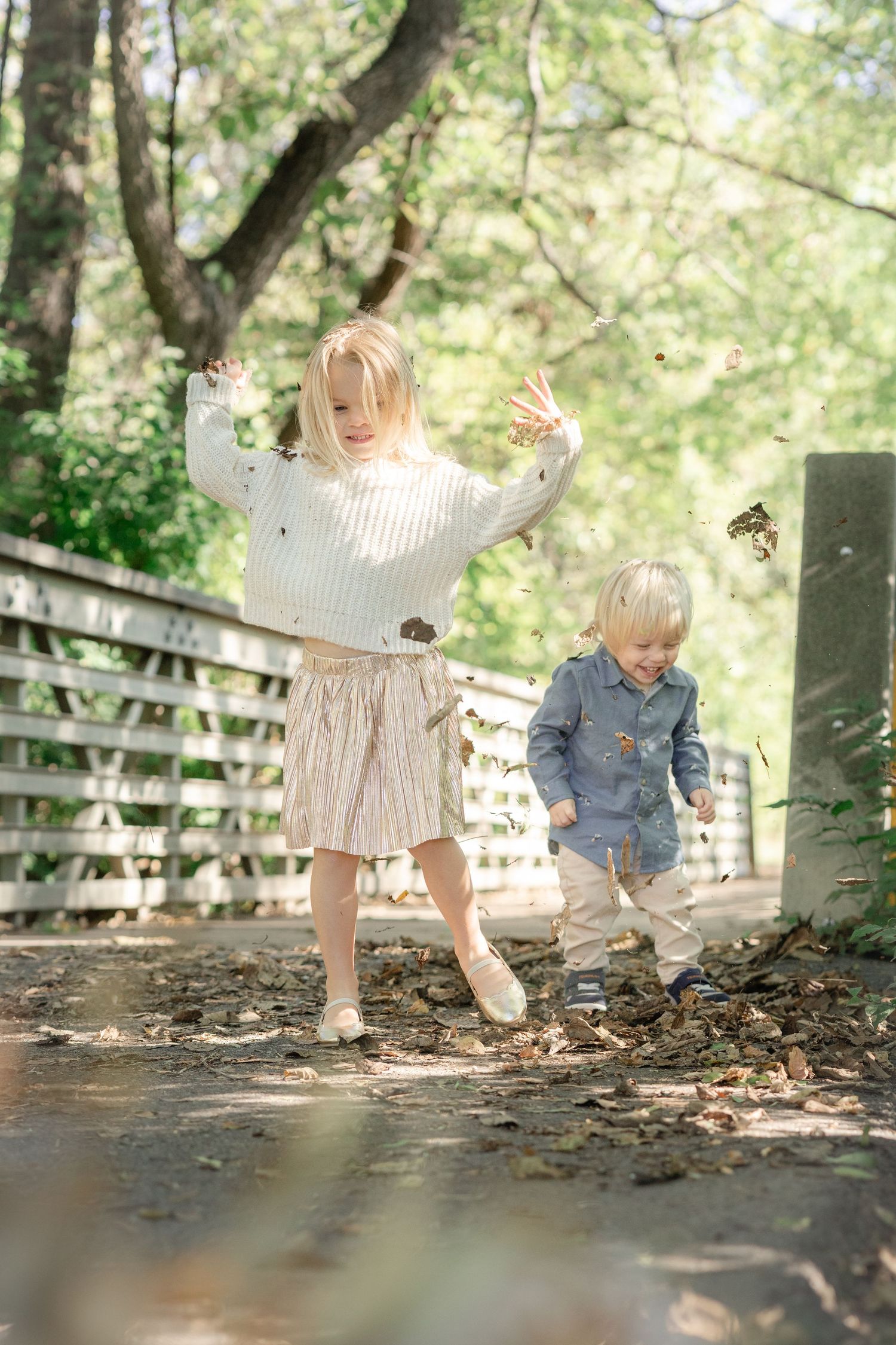Two young blonde children playfully kick fall leaves on a sunlit path near a white fence outdoors.