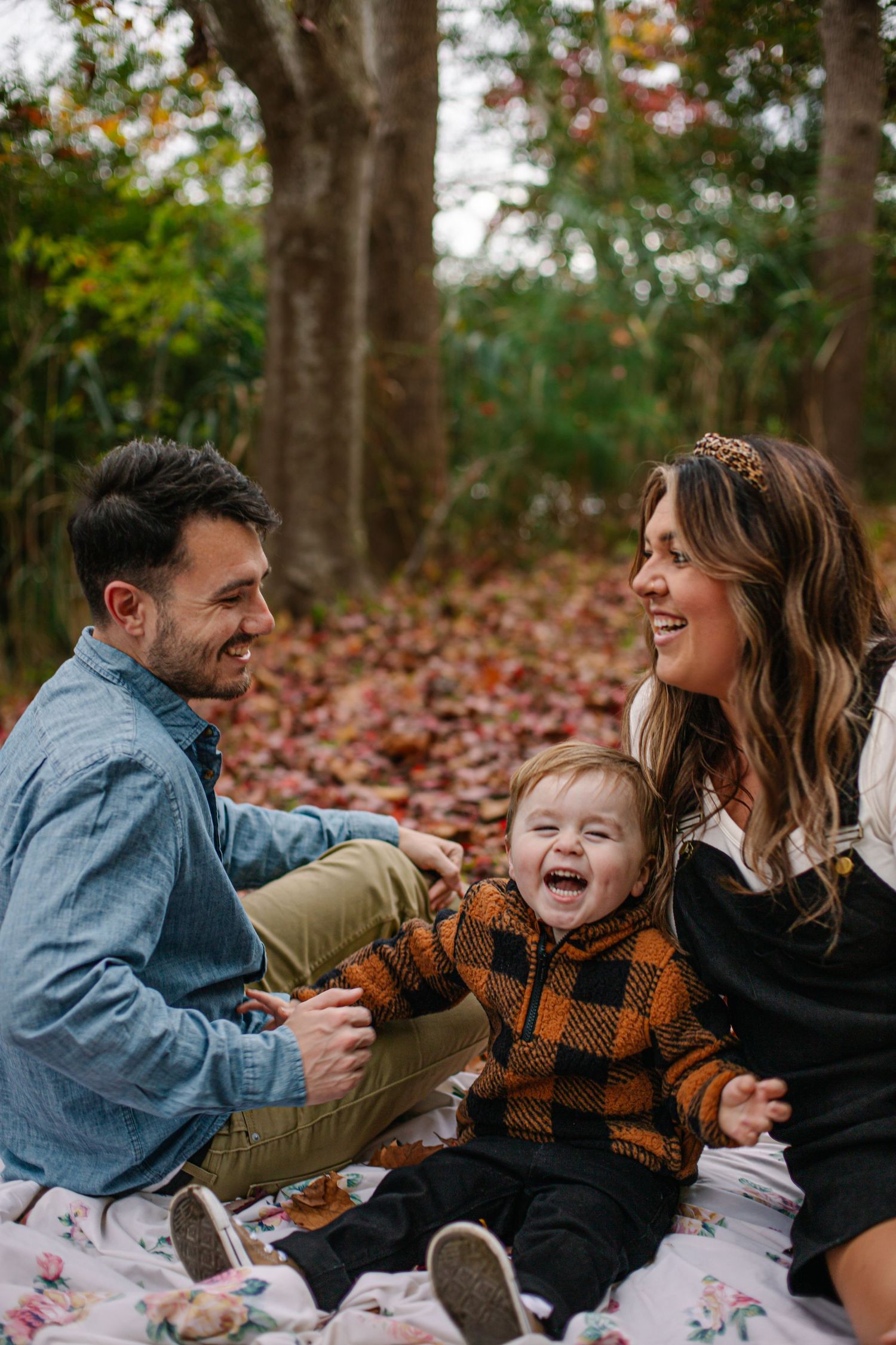 A joyful family of three sits on a floral blanket among autumn leaves, laughing together in a wooded setting.