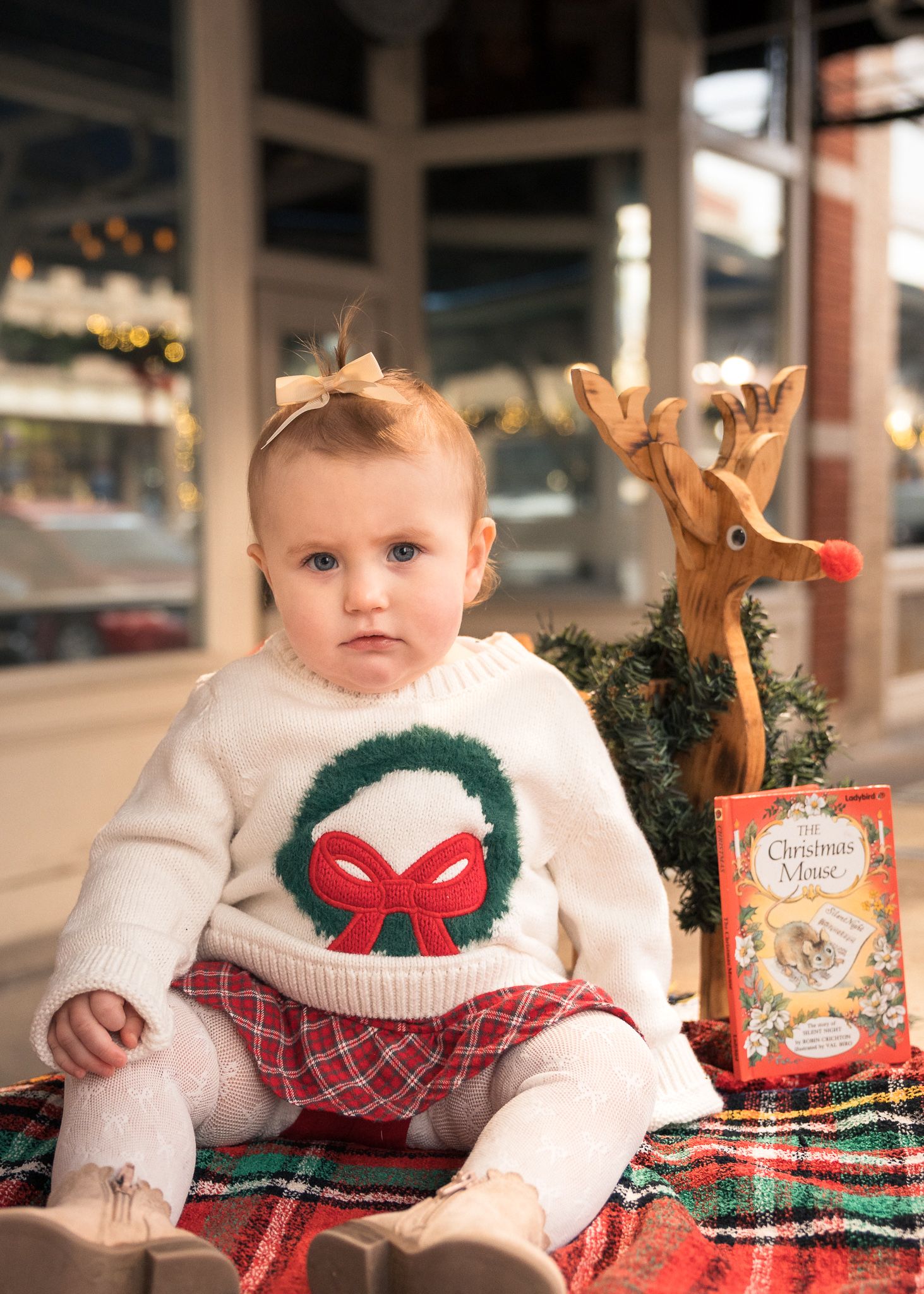 Baby in Christmas wreath sweater sits on plaid blanket with wooden reindeer and holiday book.