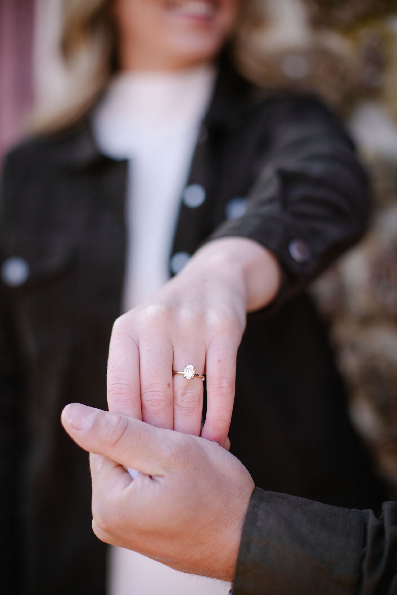 Close-up of a woman's hand displaying a gold solitaire engagement ring held by her partner outdoors.