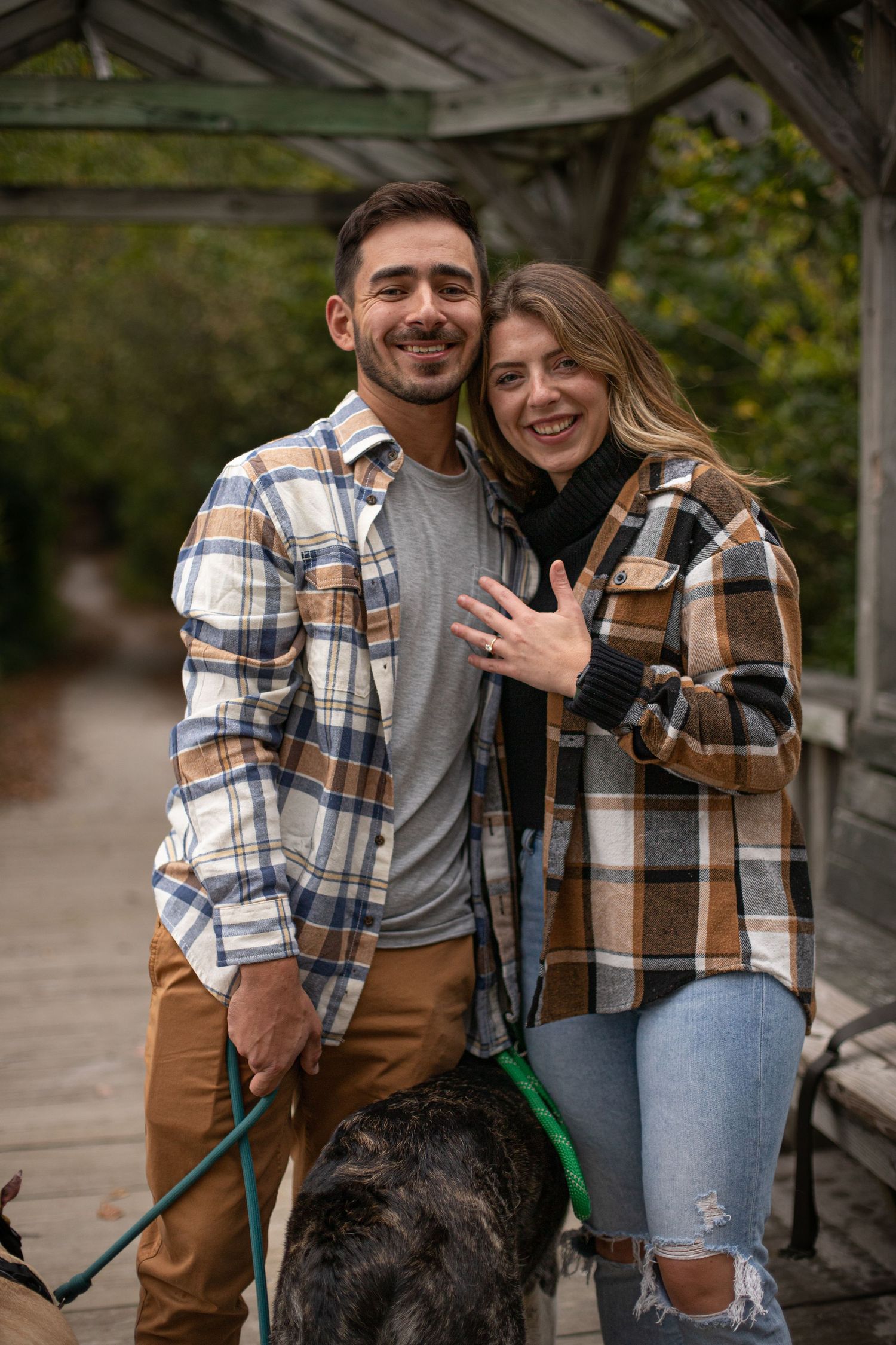 Happy couple in matching plaid shirts showing engagement ring while walking dog in park.