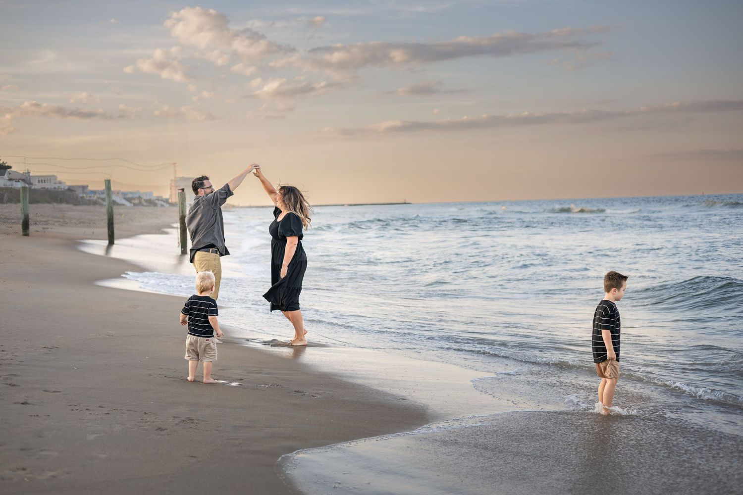 Family of four enjoying a beach sunset, parents high-fiving while two young boys play in the ocean waves.