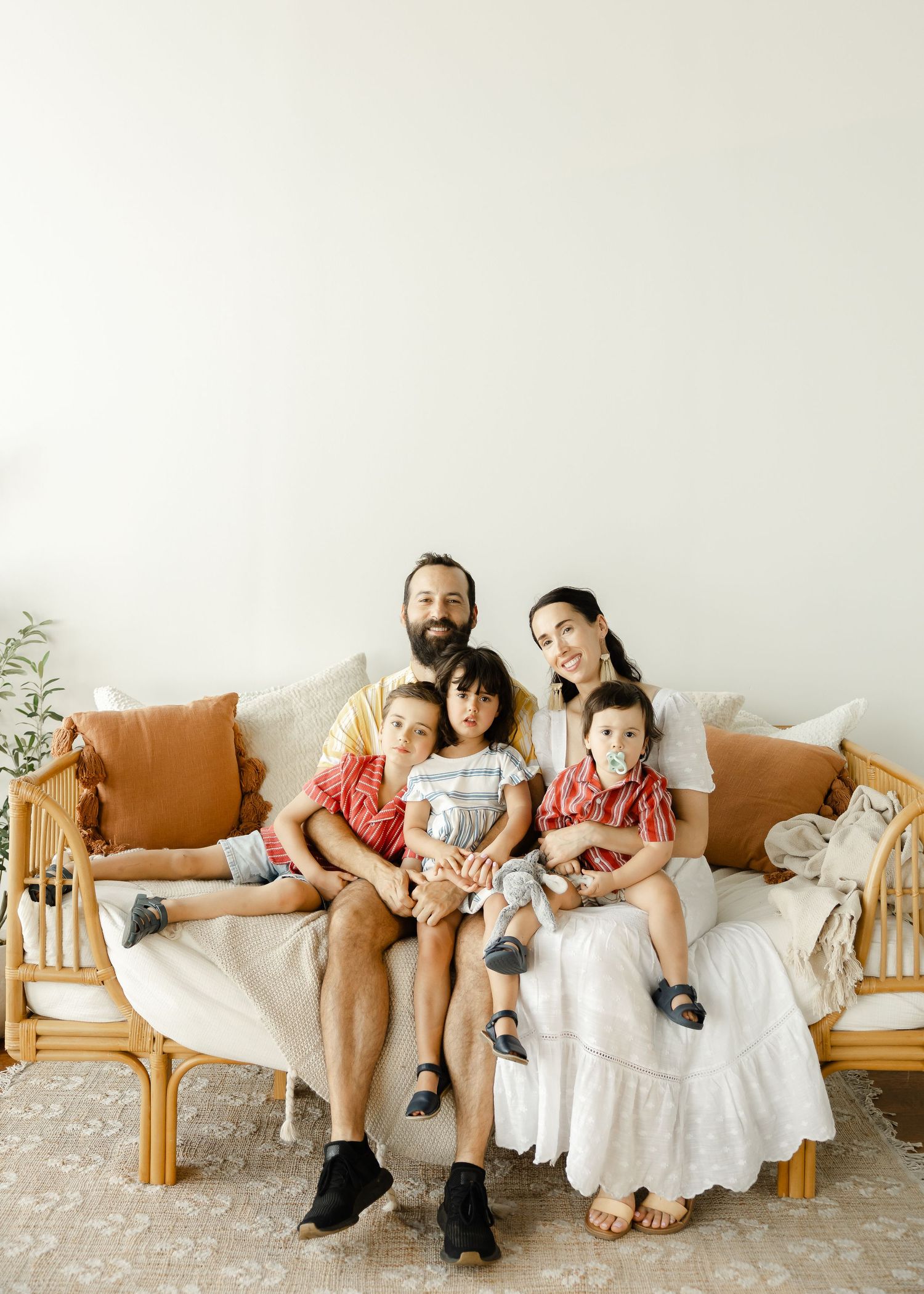 A family of five poses on a rattan sofa with tan cushions, parents flanking three young children in a bright, airy room.
