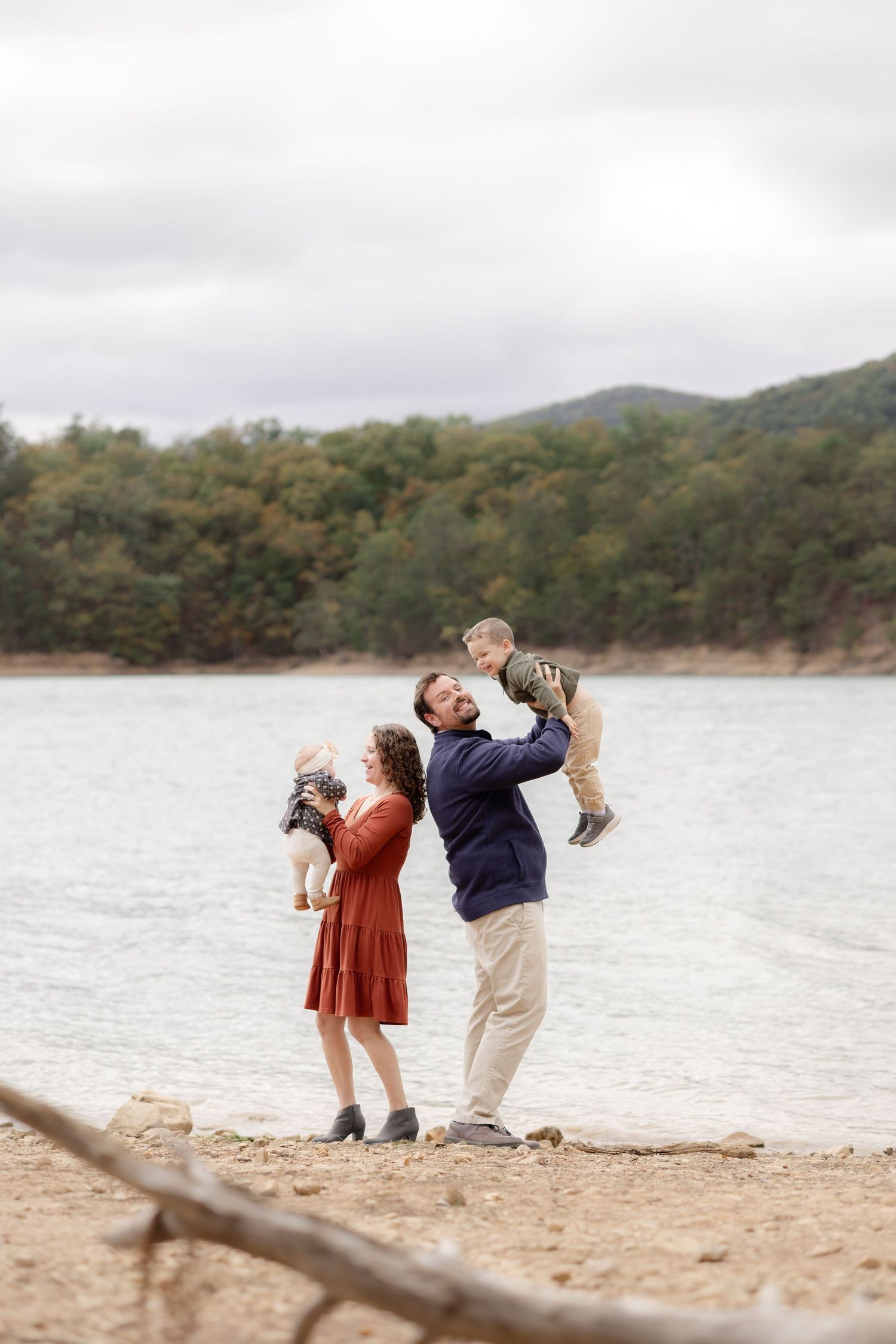 Family posing by a lake, man lifting toddler while woman holds small dog, autumn trees and cloudy sky in background. Roanoke, VA