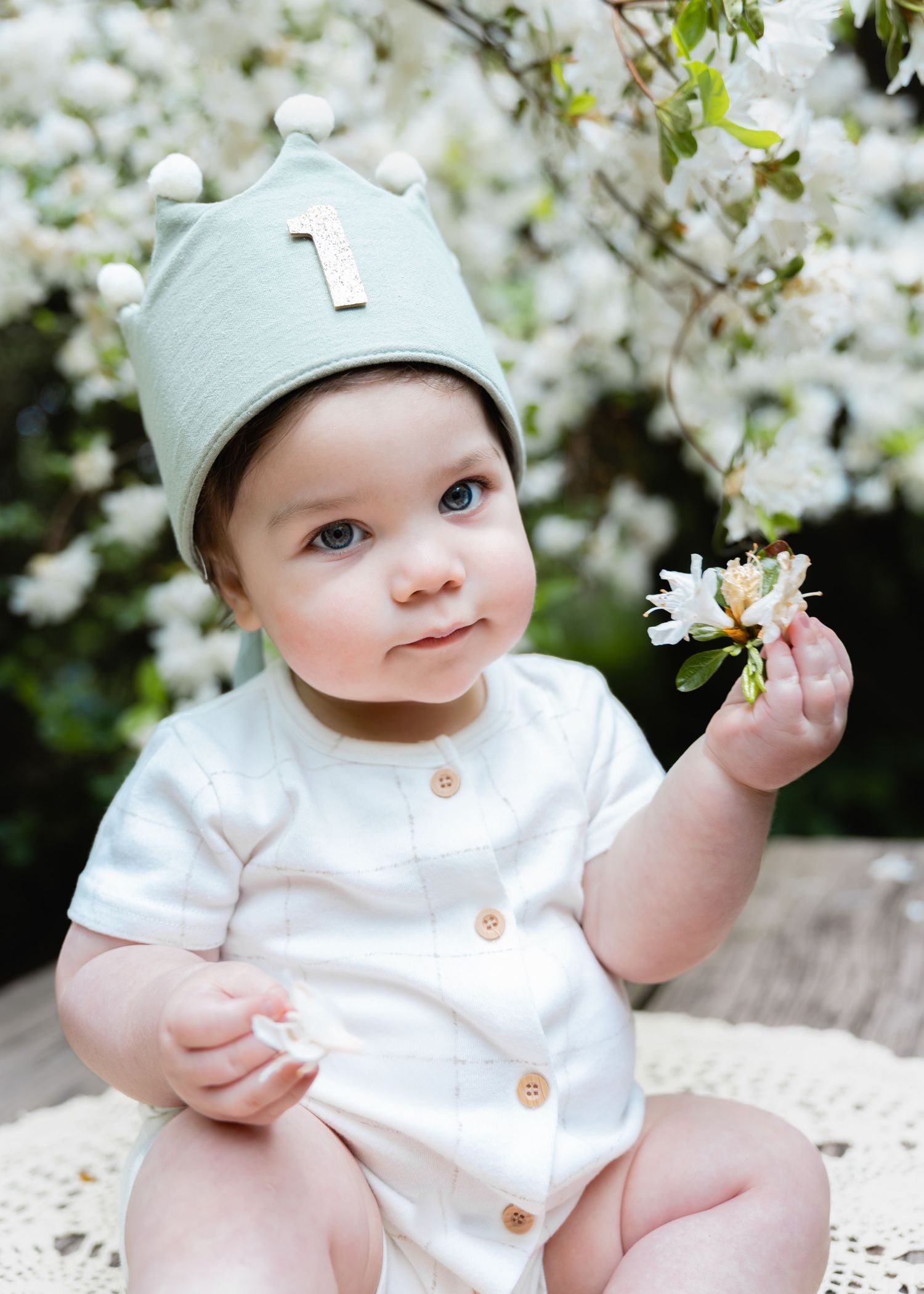 Baby wearing green crown hat holds a flower, sitting outdoors among white blossoms, celebrating first birthday.