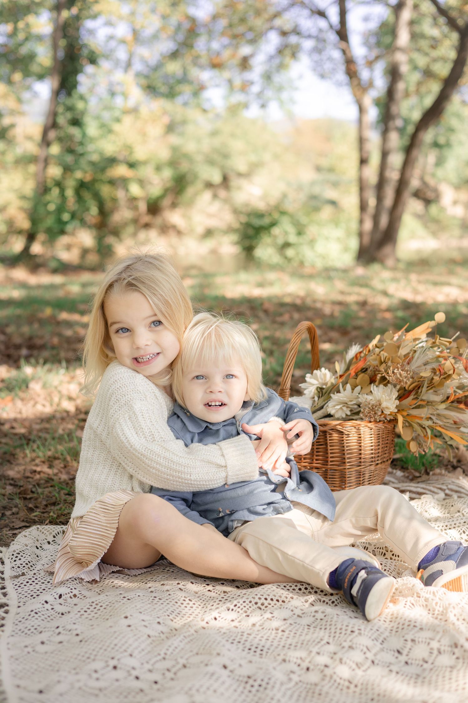 Two blonde siblings hug on a lace blanket during an autumn picnic with a wicker basket of fall flowers.