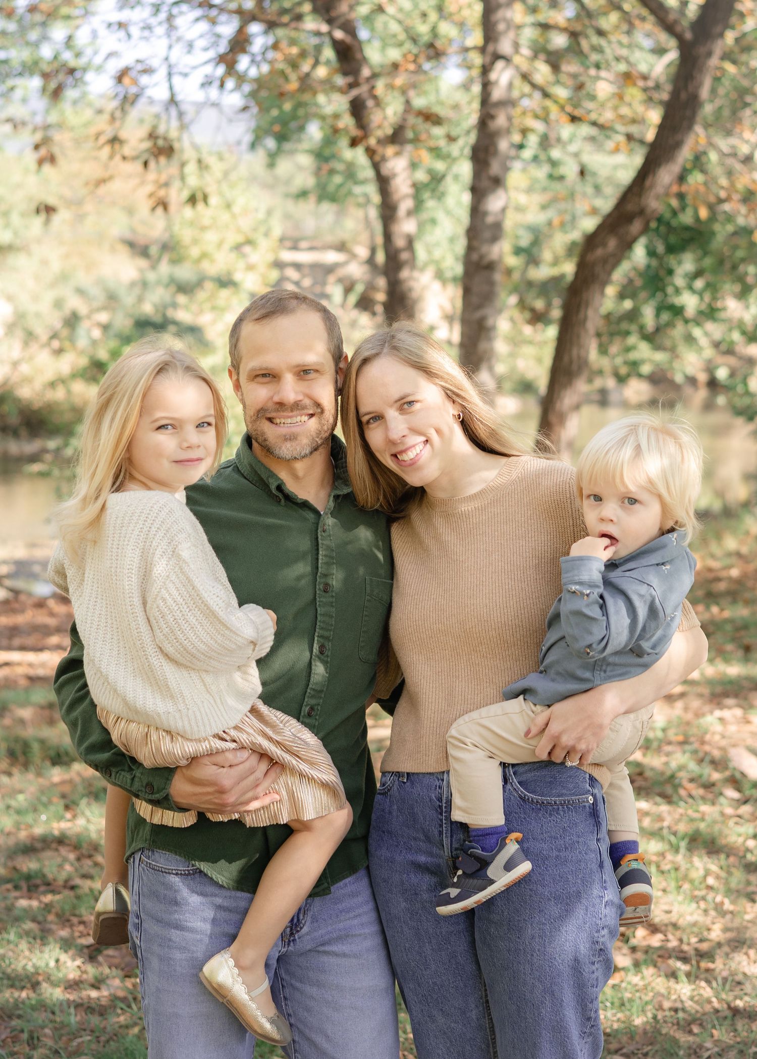 A smiling family of four poses outdoors in autumn, parents holding two young children among colorful trees.