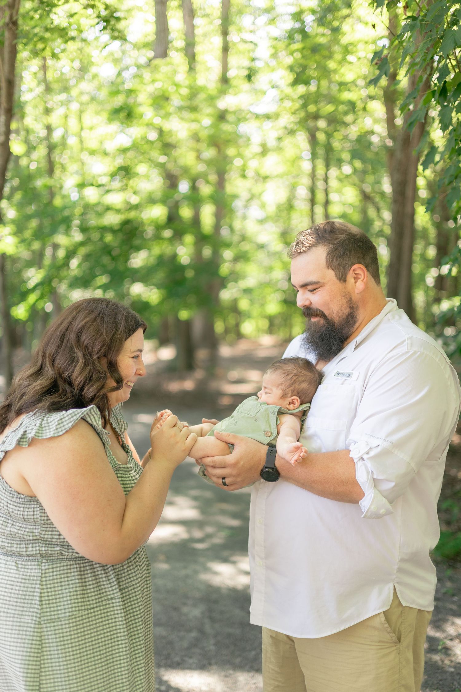 A couple stands in a lush green forest path, smiling at their newborn baby held in the fathers arms.