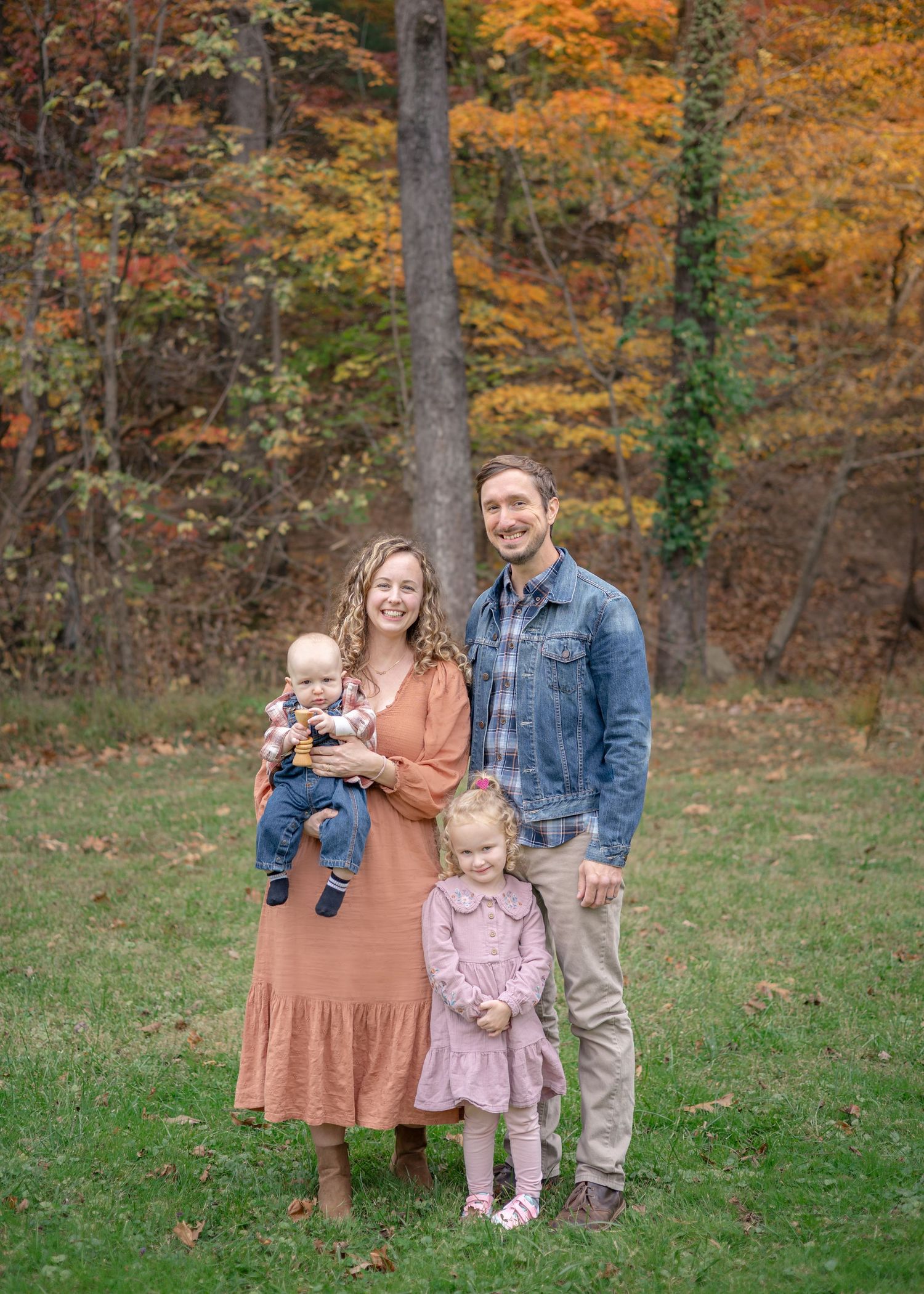 Family of four posing outdoors in autumn, mother holding baby, toddler girl standing, colorful fall foliage in background.