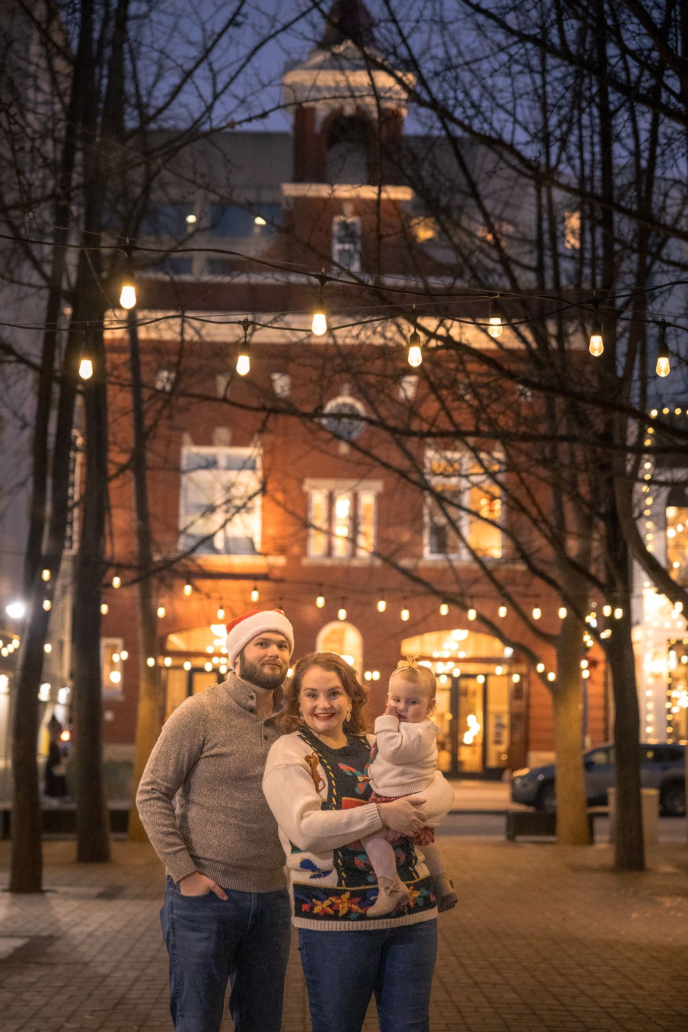 Family posing outdoors at night with festive string lights and brick building during Christmas season.