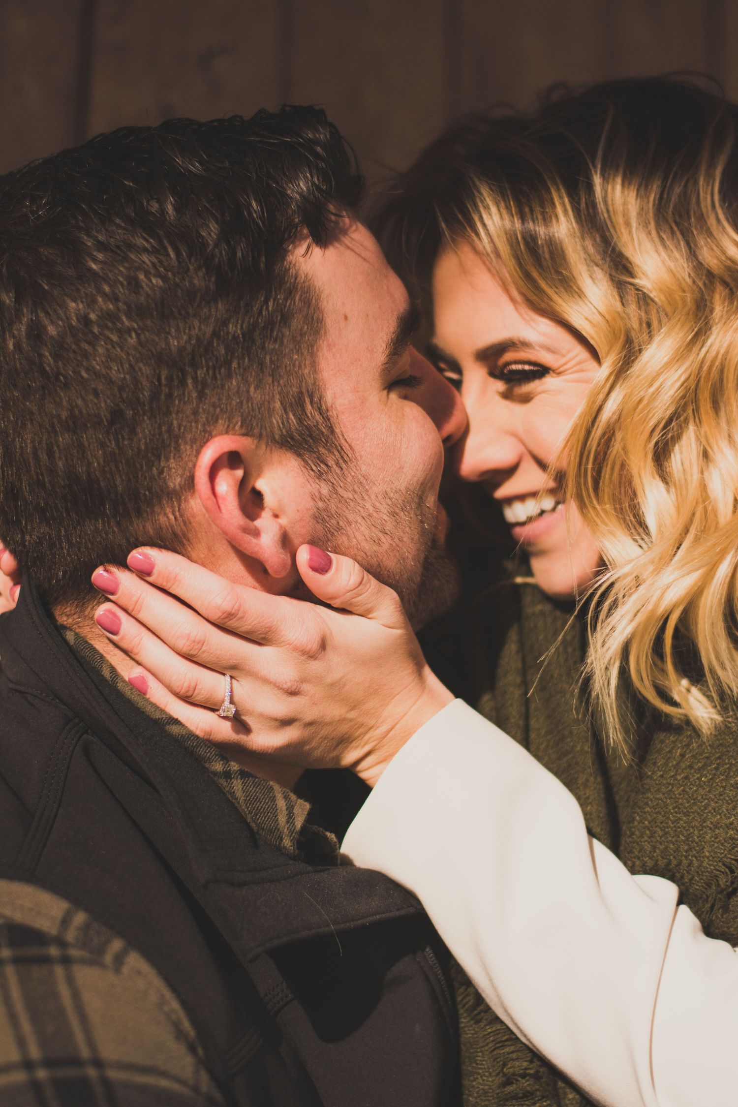 A smiling blonde woman with pink nails holds a man's face close to hers in a romantic moment, wearing an engagement ring.