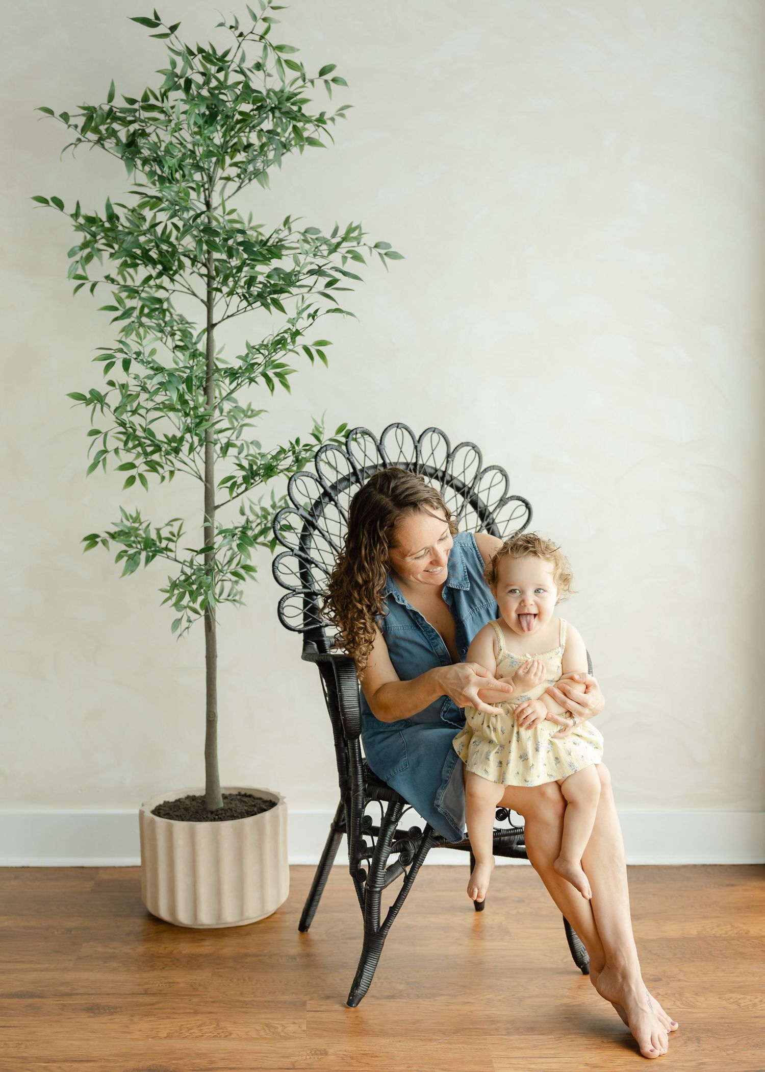 A mother and toddler laugh together in a black rattan chair beside a potted tree on a wood floor.
