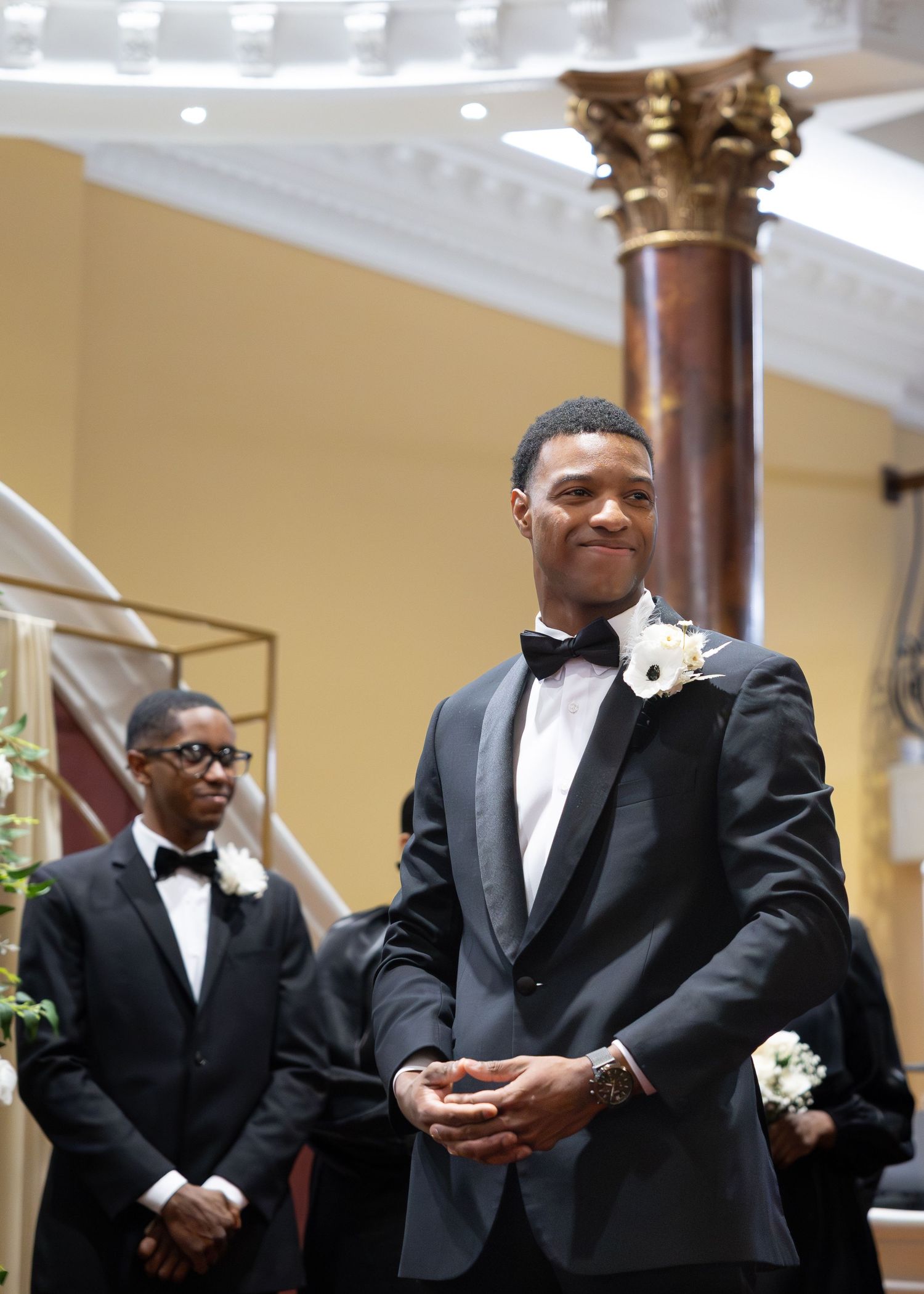 Groom in black tuxedo with white boutonniere smiling at wedding ceremony.