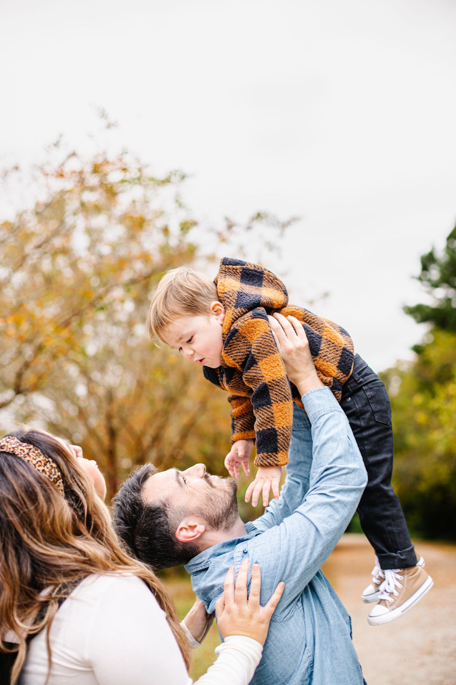 Family enjoying autumn outdoors, father lifting toddler in plaid jacket while mother watches and smiles in a park setting.