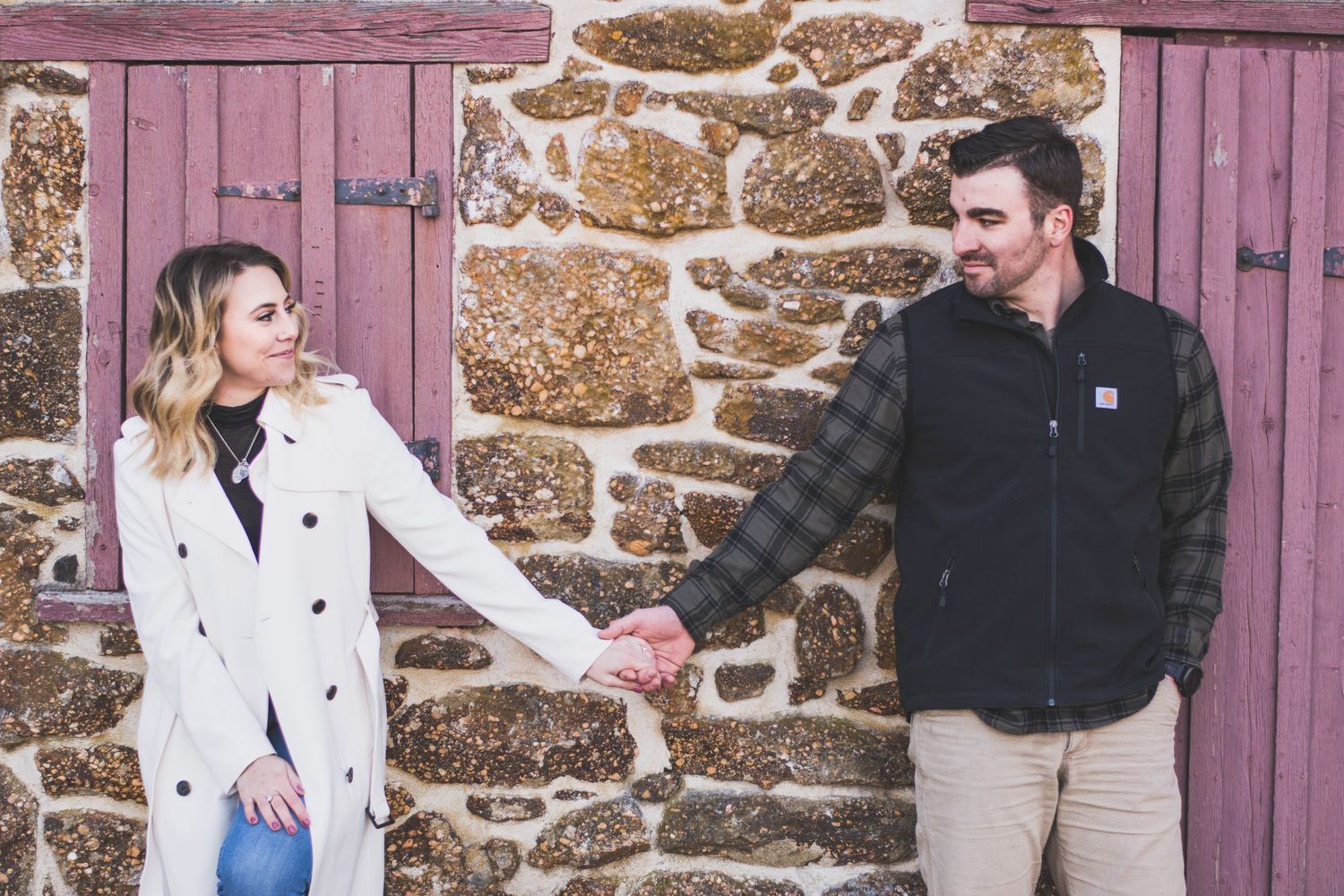 Couple holding hands in front of a rustic stone wall with wooden doors during an outdoor engagement photo session.