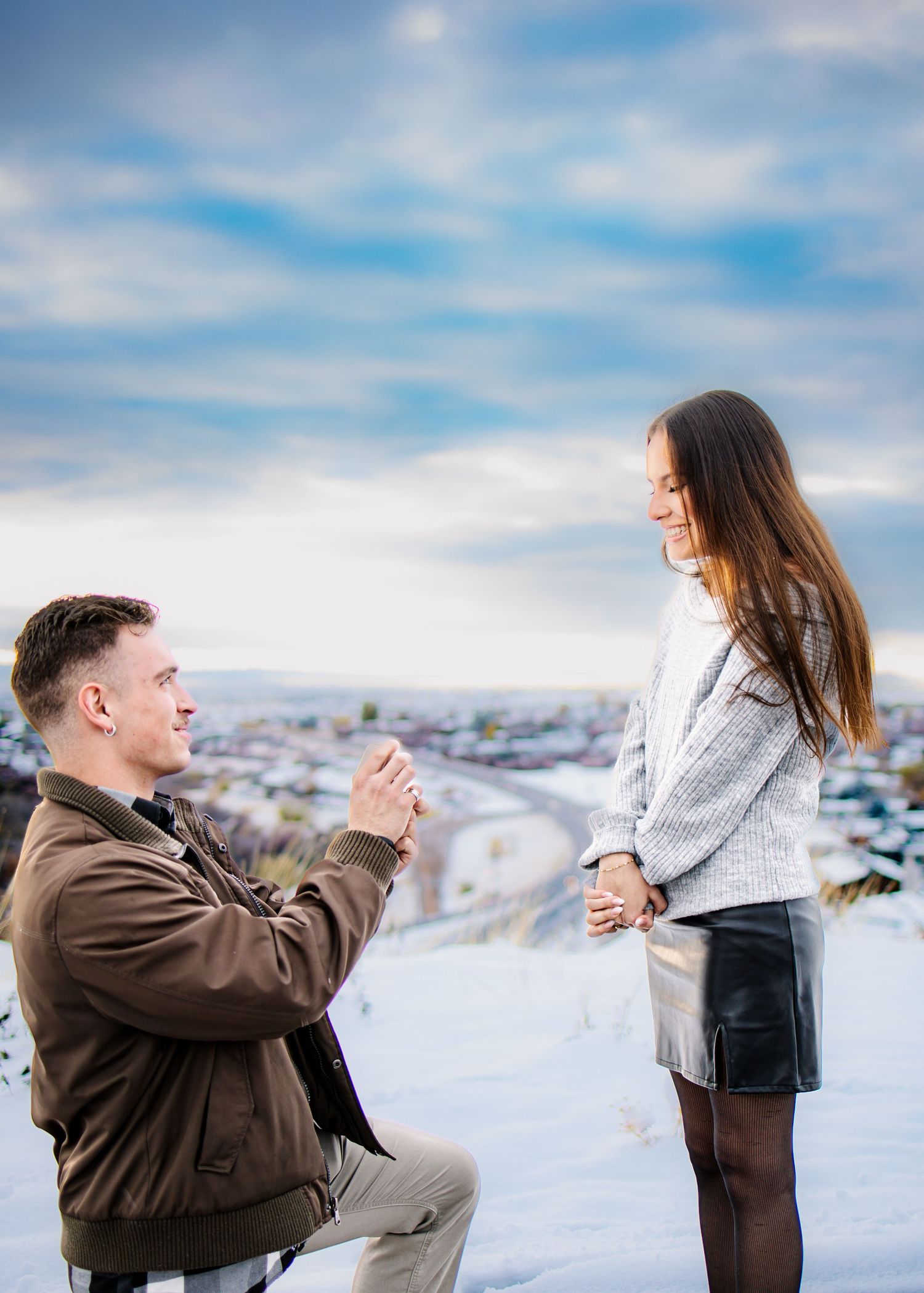 Man proposing on one knee to woman on snowy hilltop overlooking a city under a dramatic winter sky.