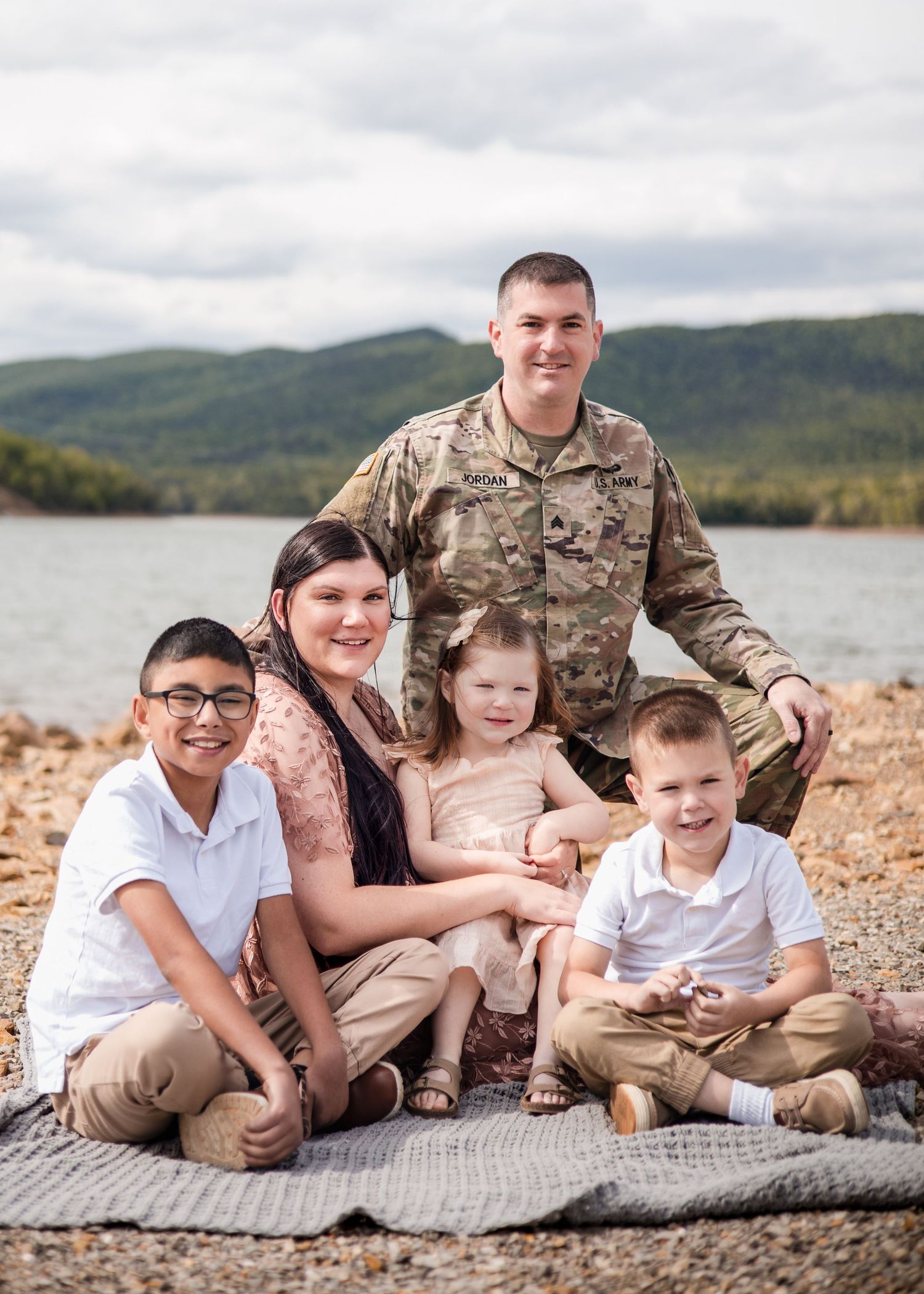 A military family of five poses outdoors by a lake with green hills, featuring a soldier in camo uniform with his wife and three children.