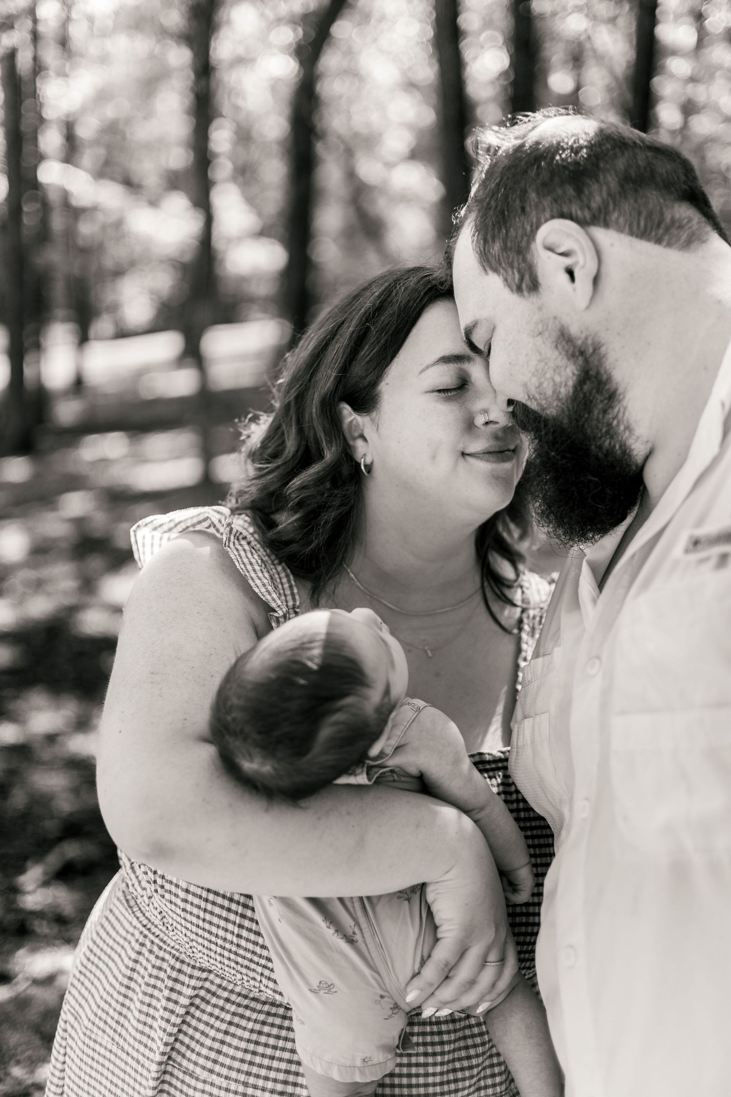 Black and white photo of a couple sharing a tender forehead touch while holding their newborn baby in a forest setting.