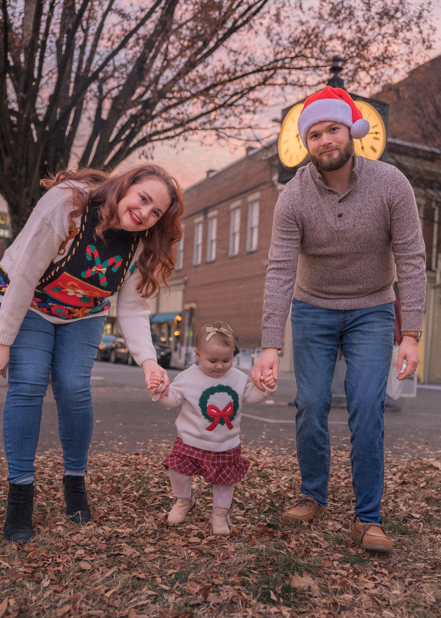 Family of three posing outdoors in Christmas sweaters, parents holding toddler's hands on fallen leaves at dusk.