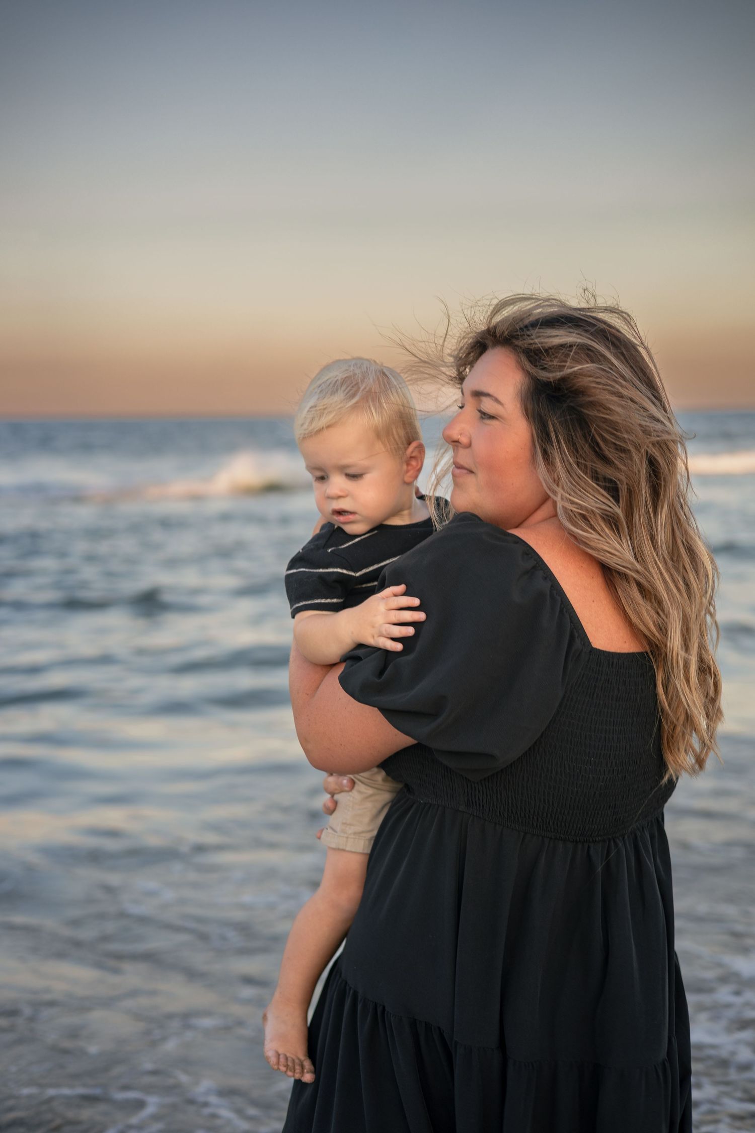 A mother in a black dress holds her toddler son on a beach at sunset with ocean waves behind them.