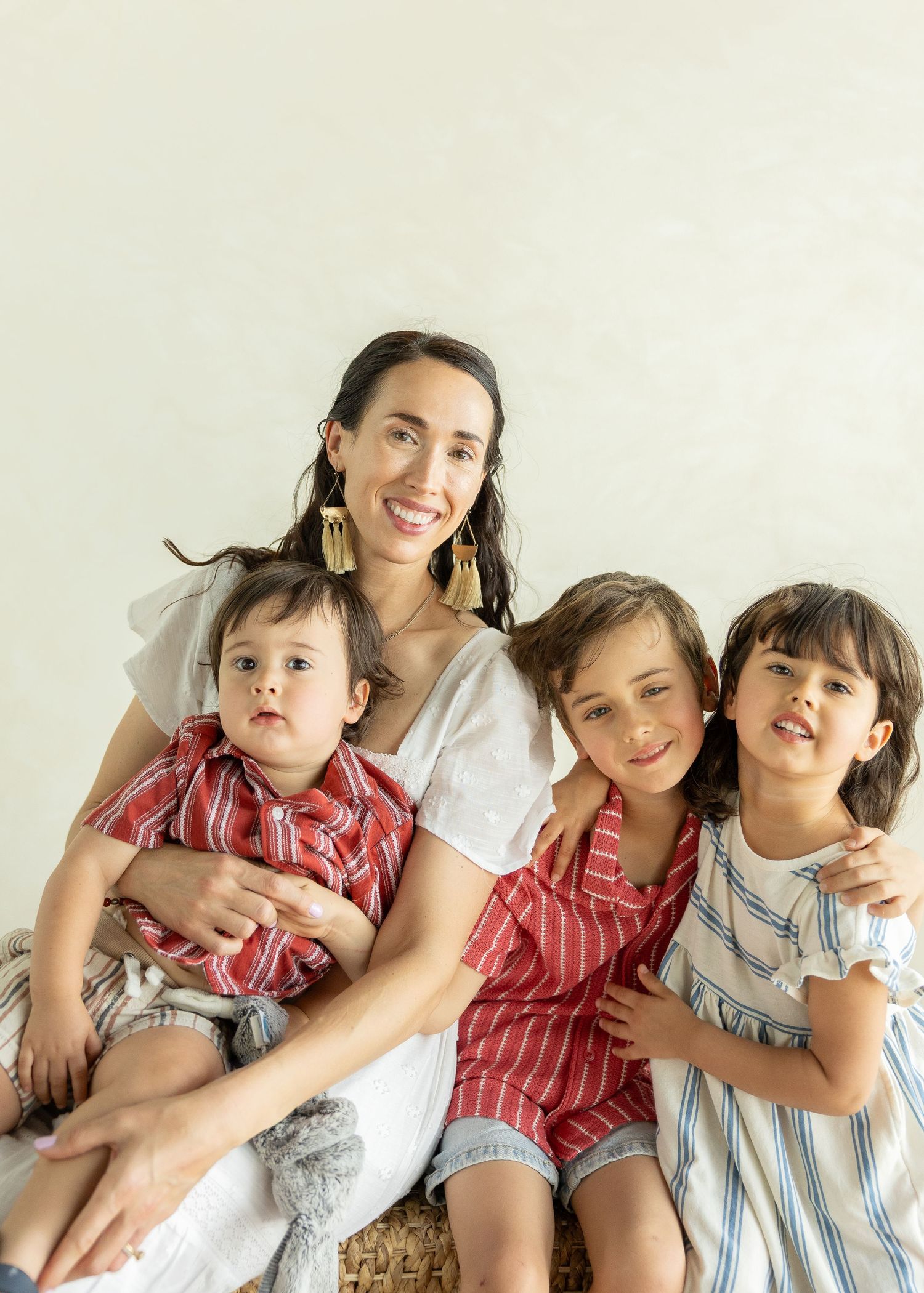 A smiling woman with gold earrings holds a baby boy while two young children lean in for a bright family portrait.