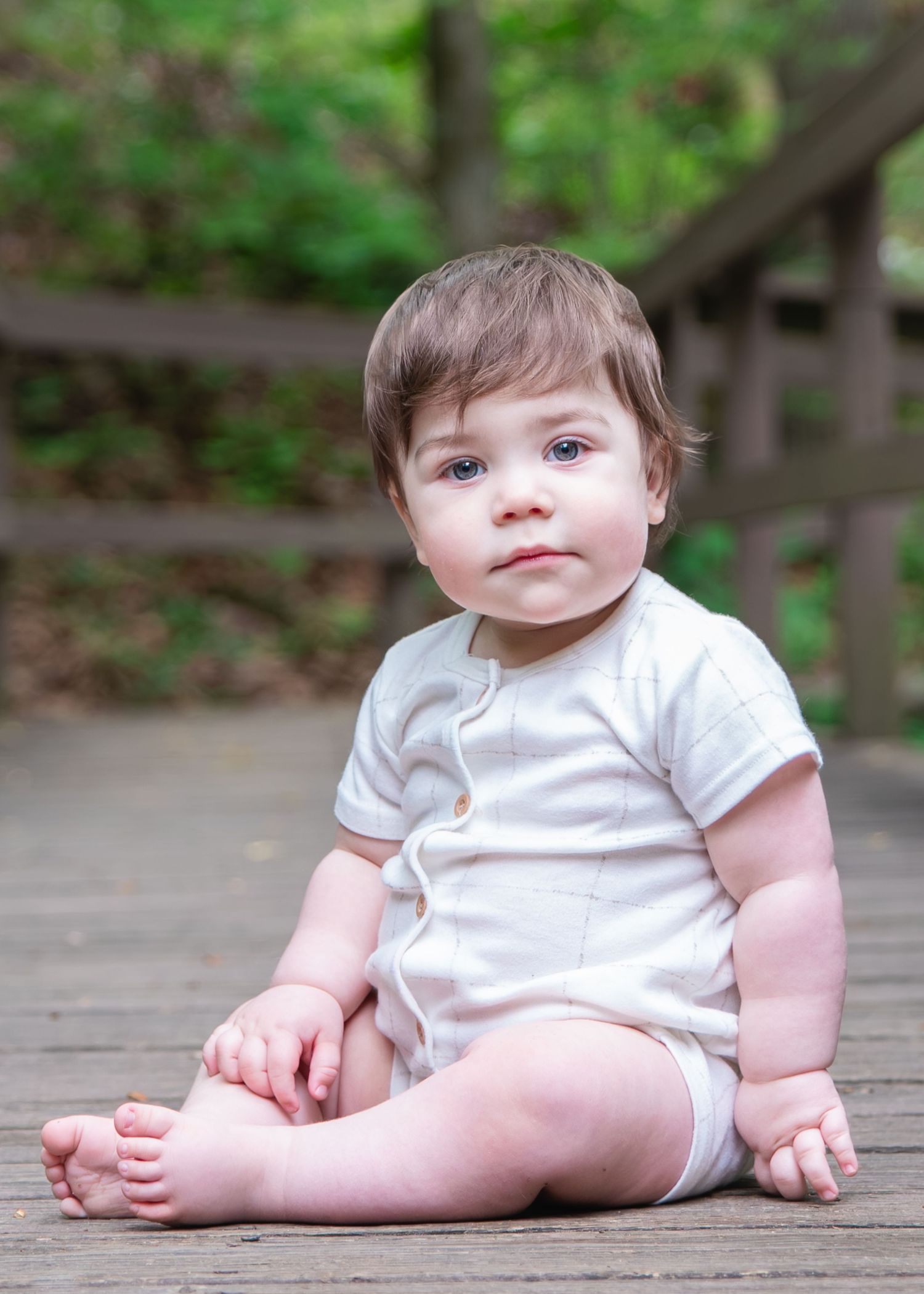 Adorable baby in white onesie sitting on wooden deck outdoors surrounded by lush green trees.