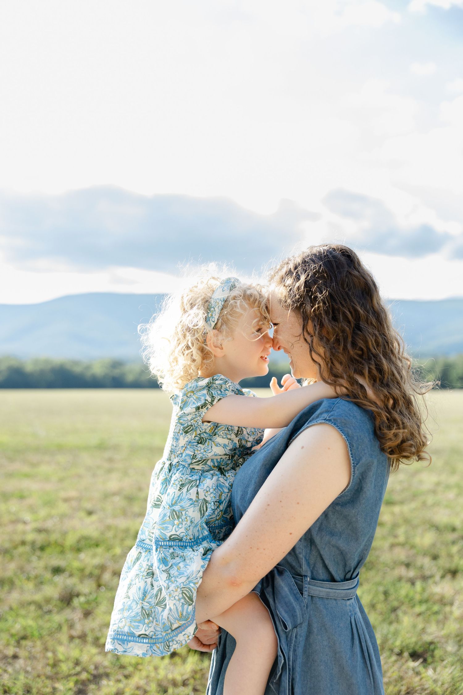A mother in a blue dress holds her curly-haired toddler girl as they touch foreheads in a green field.