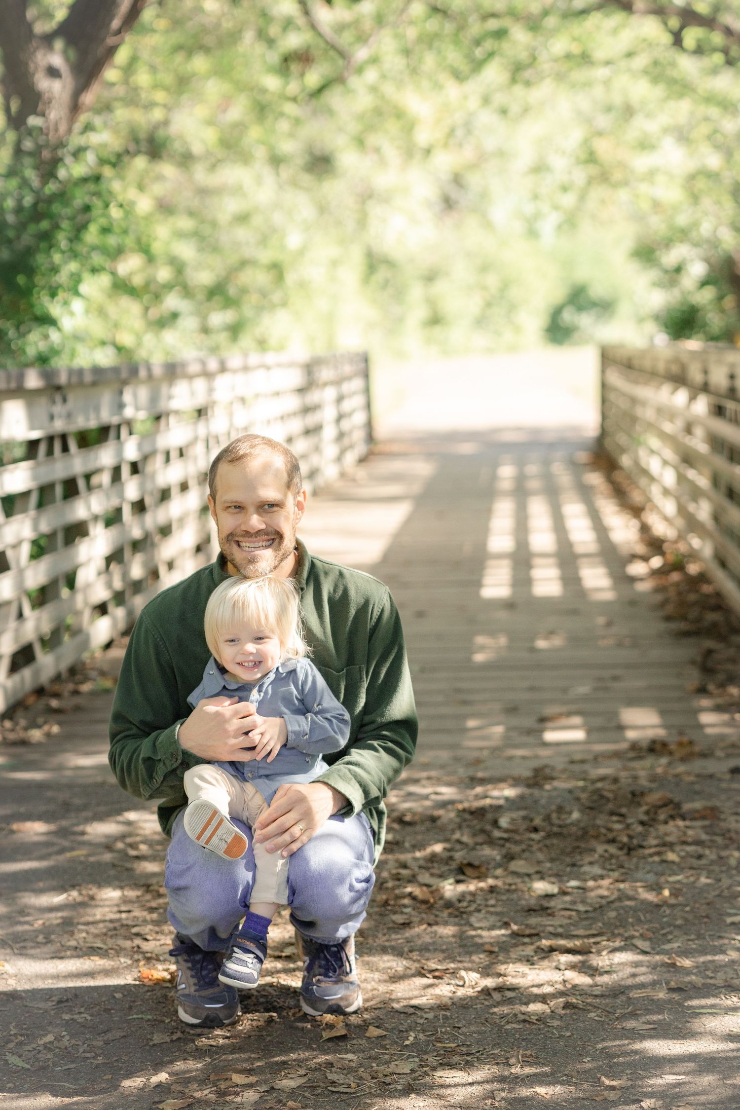 Father crouching with toddler on a sunlit wooden bridge path surrounded by green trees.