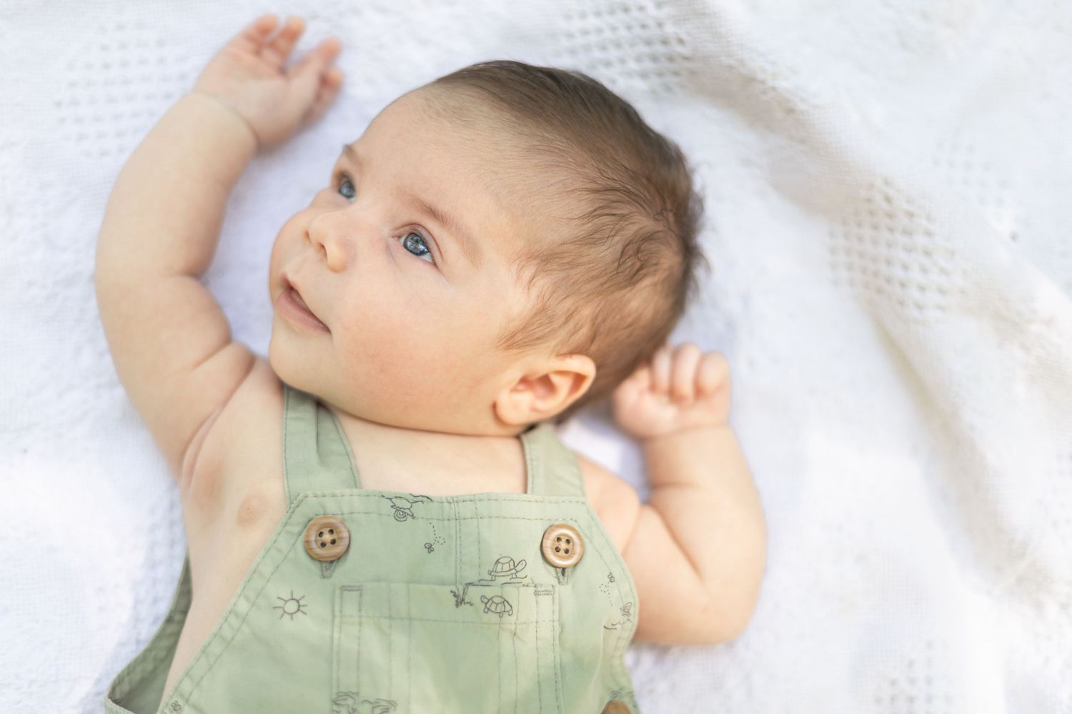 Newborn baby in green overalls lying on white blanket, looking up with blue eyes and raised arms.