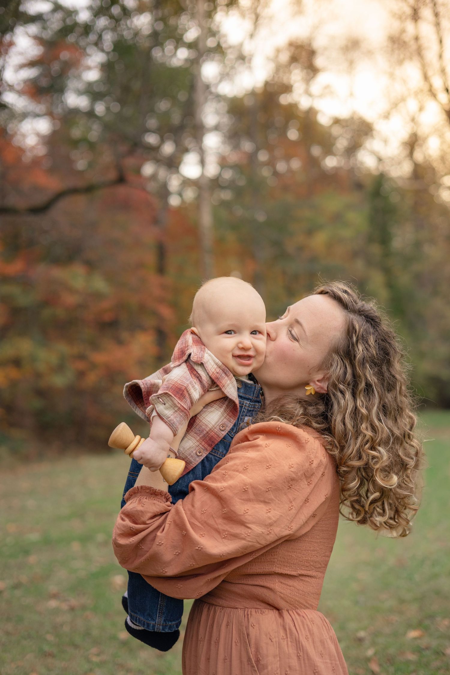 A mother with curly hair kisses her smiling baby outdoors in an autumn park setting.