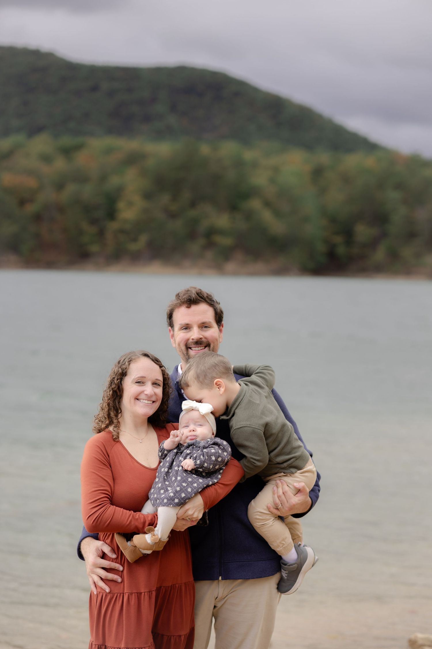 Family of four poses by a lakeside with forested mountains, parents holding two young children on a cloudy day. Carvin's cove, roanoke, va